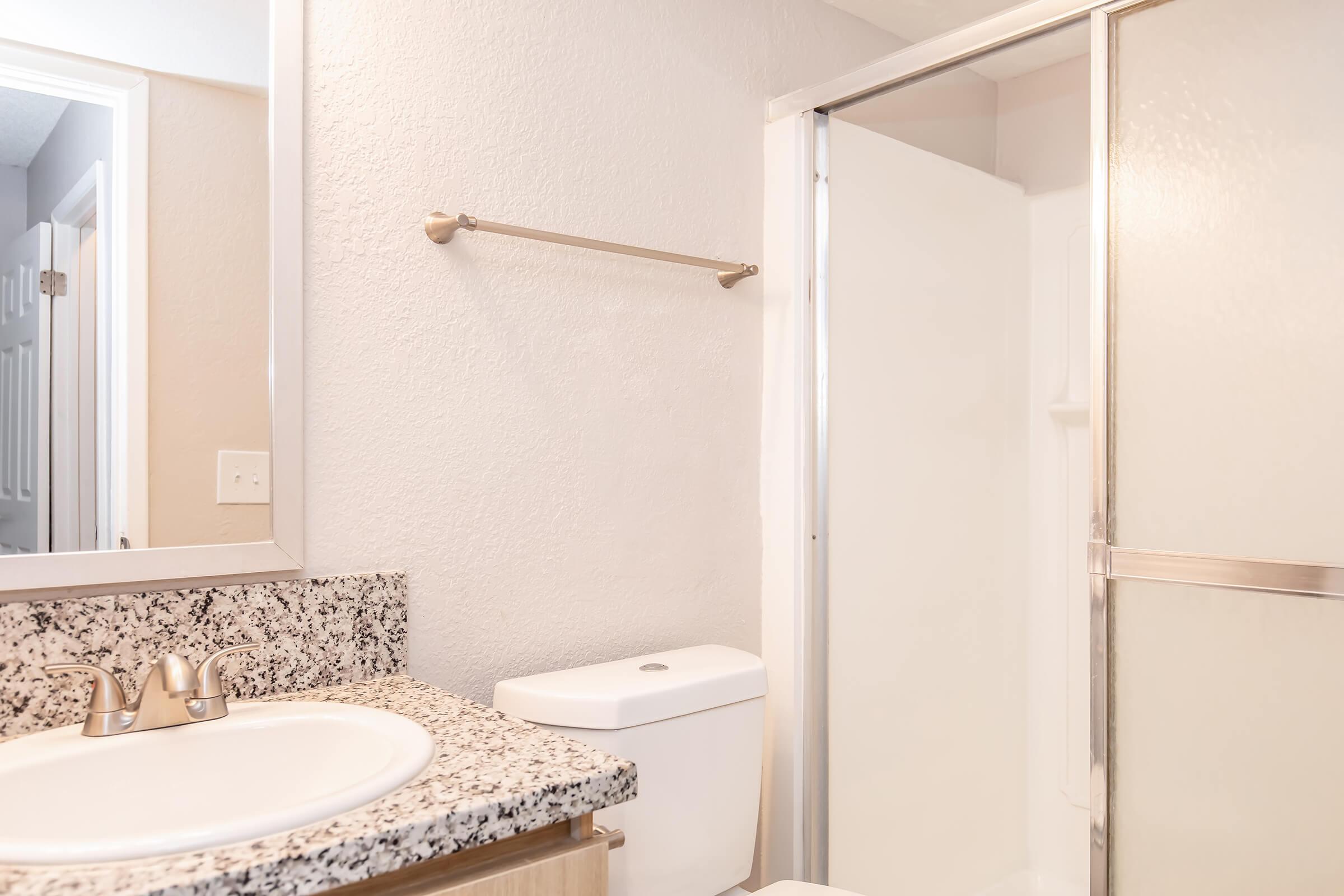 A clean and modern bathroom featuring a white sink with a granite countertop, a toilet, and a clear glass shower enclosure. Light-colored walls and good lighting create a bright atmosphere, while a towel bar is mounted on the wall. A mirror above the sink completes the space.