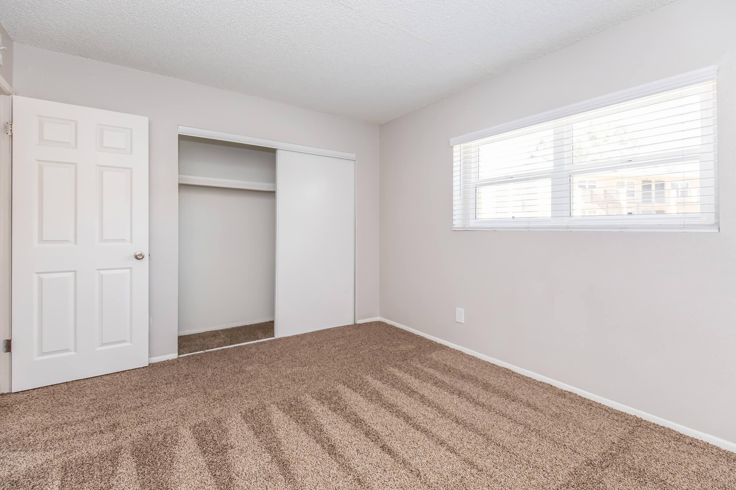 A cozy, empty room with light beige walls and a large window. The floor is covered in light brown carpet, and there is a closet with sliding doors on the left. The room appears bright and airy, with natural light illuminating the space.