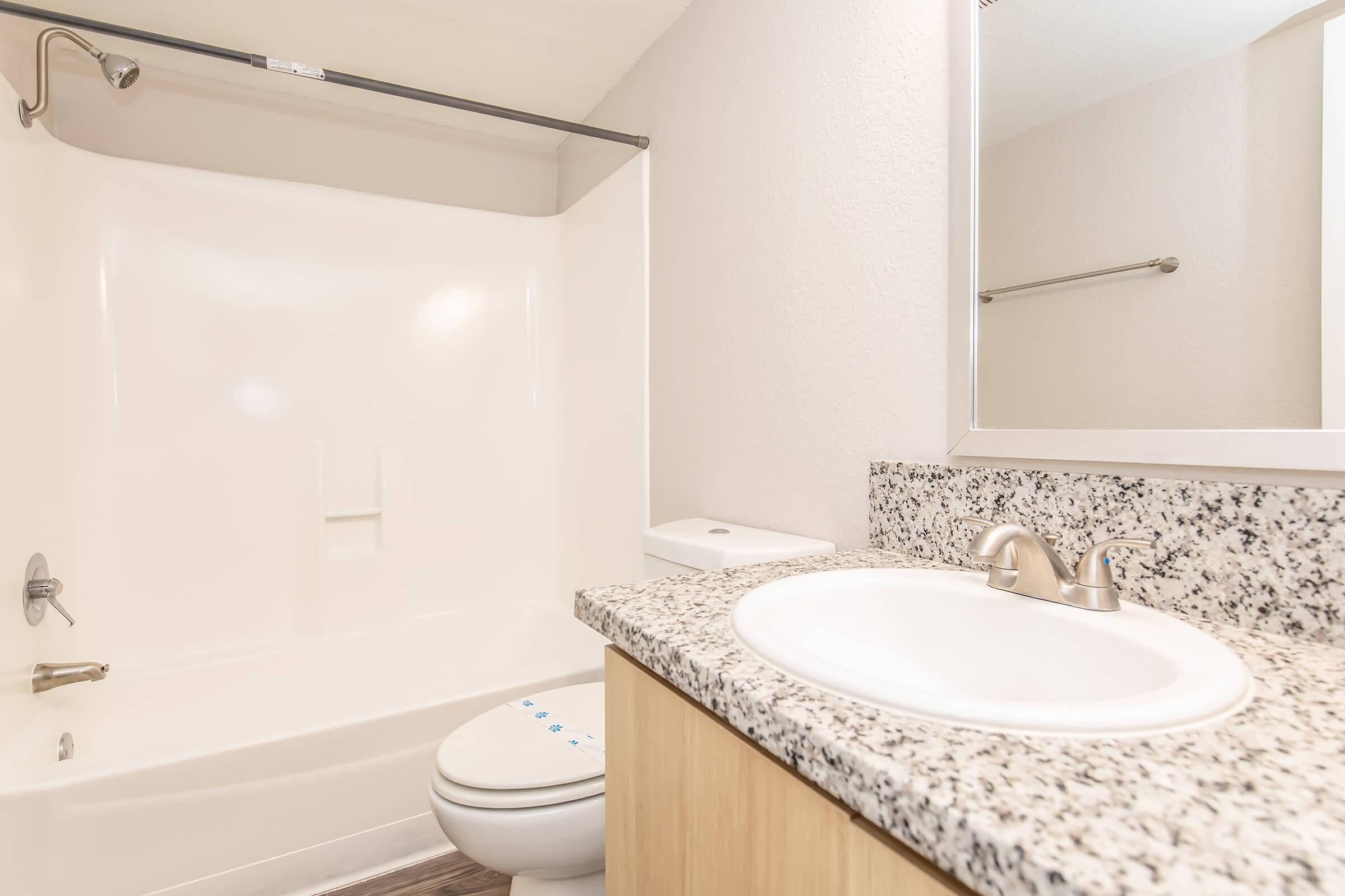 A clean and modern bathroom featuring a bathtub with a shower, a light-colored granite countertop with a sink, and a mirror above the sink. A towel rack is mounted on the wall, and the overall color scheme is neutral and inviting, creating a spacious feel.