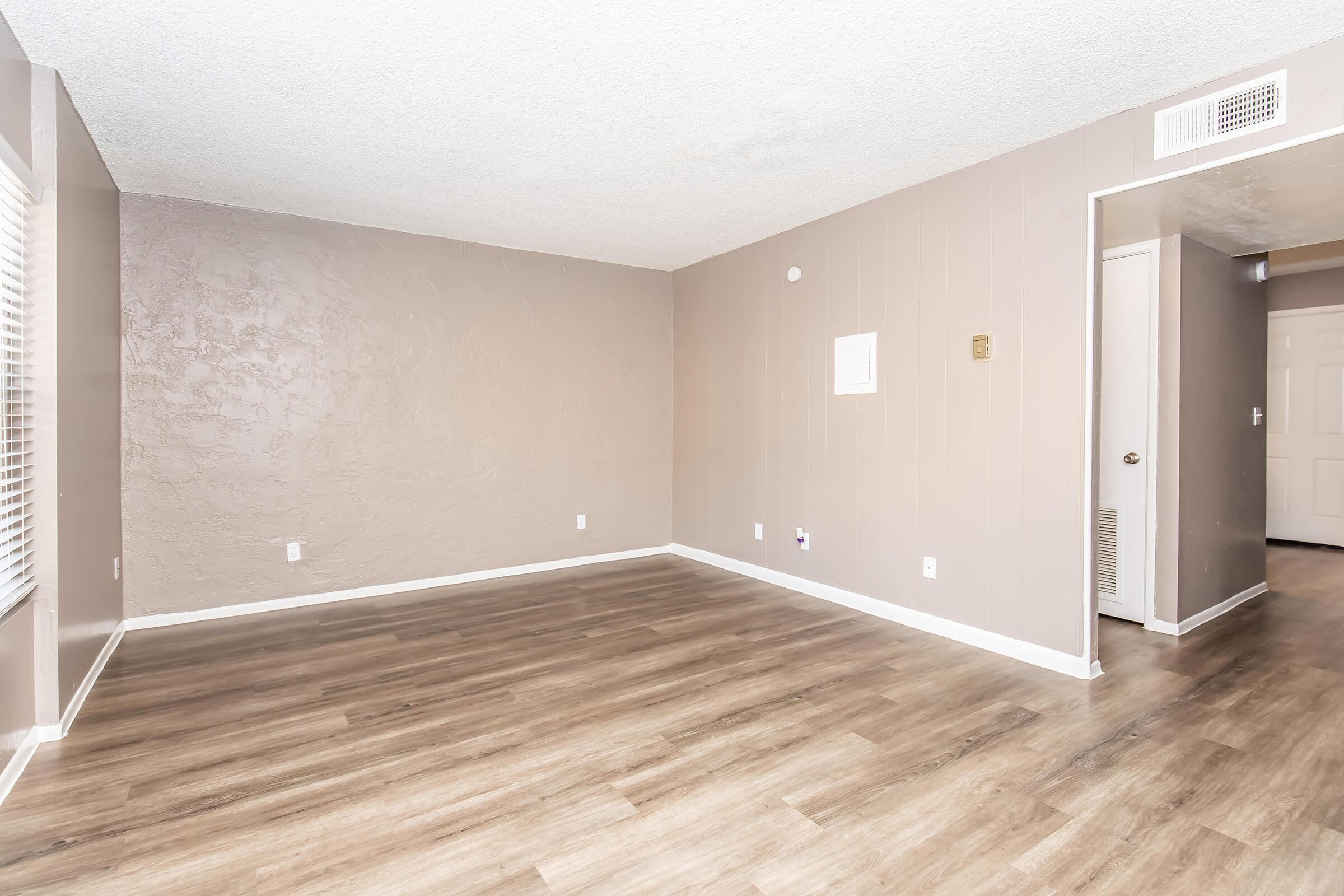 Empty living room with light brown walls and new laminate flooring. A window with blinds on the left side and a doorway leading to another room on the right. The space is bright and uncluttered, featuring a minimalist aesthetic.