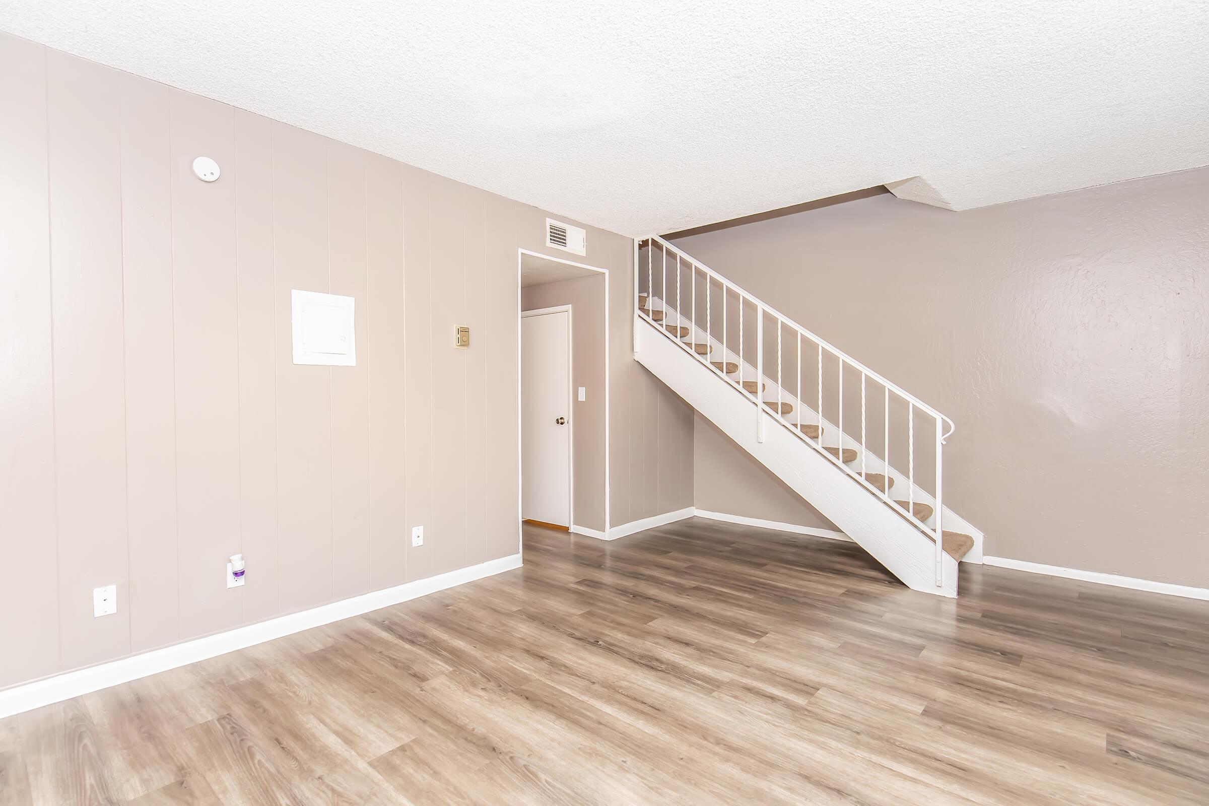 A spacious, light-filled living area featuring light brown walls and new wood-like flooring. A staircase with white railings leads to the upper floor. The room is empty, creating a clean and open atmosphere, with a small wall-mounted panel on one side. A door and light switch are visible.