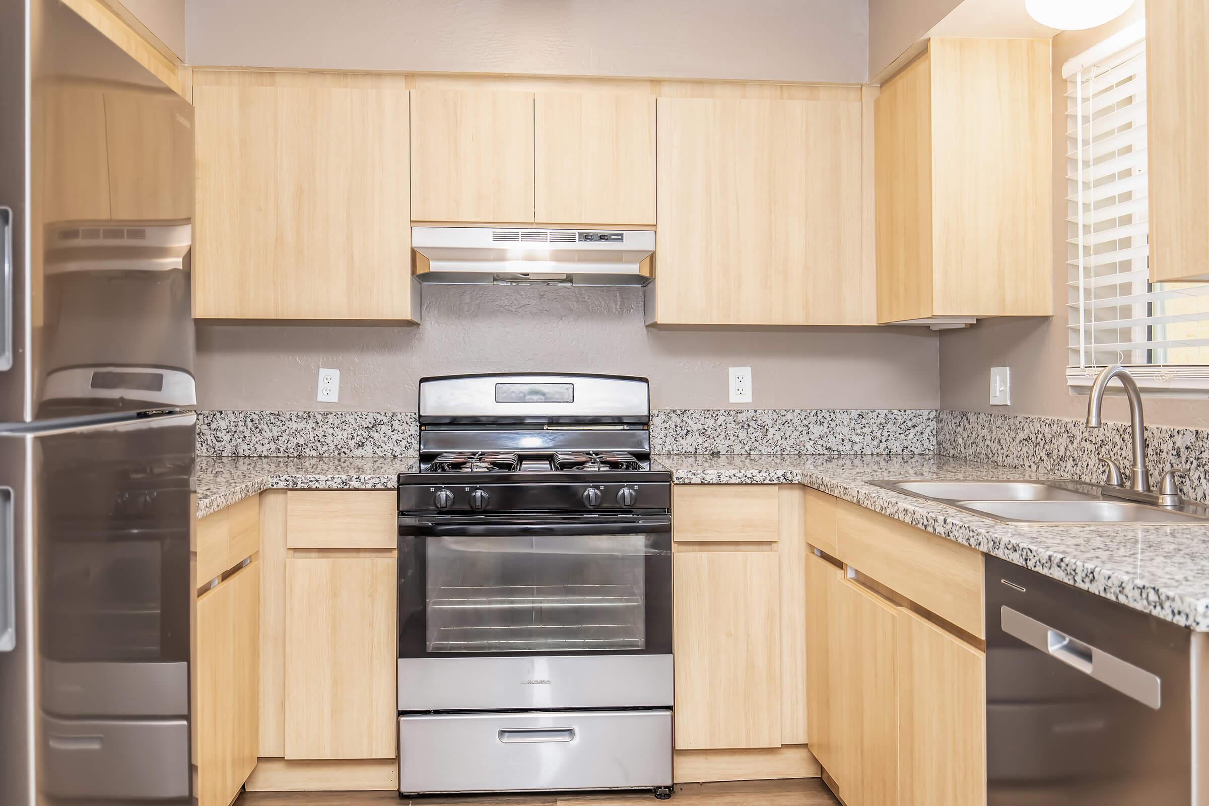 A modern kitchen featuring light wood cabinetry, a silver gas stove, and a granite countertop. The kitchen also includes a stainless steel refrigerator and a double sink, with natural light streaming in through a small window. The contemporary design highlights functionality and clean aesthetics.