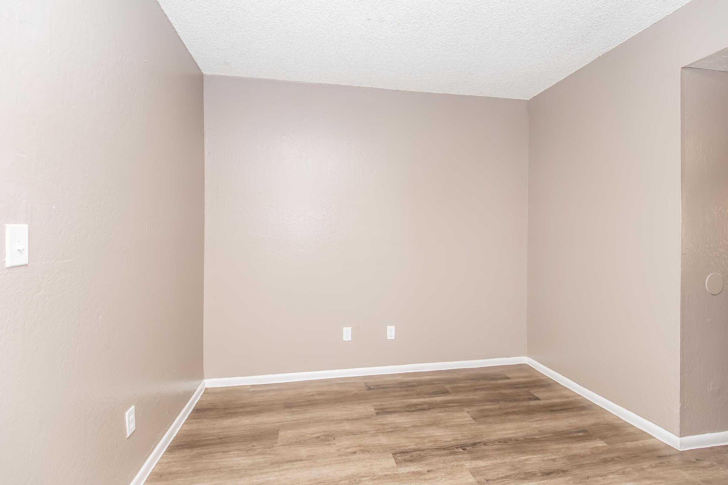 Empty room with light beige walls, a textured ceiling, and wooden flooring. The space features two electrical outlets on the wall and a small cutout or recess on the right side. The room is well-lit and has a minimalistic design, creating an open and airy feel.