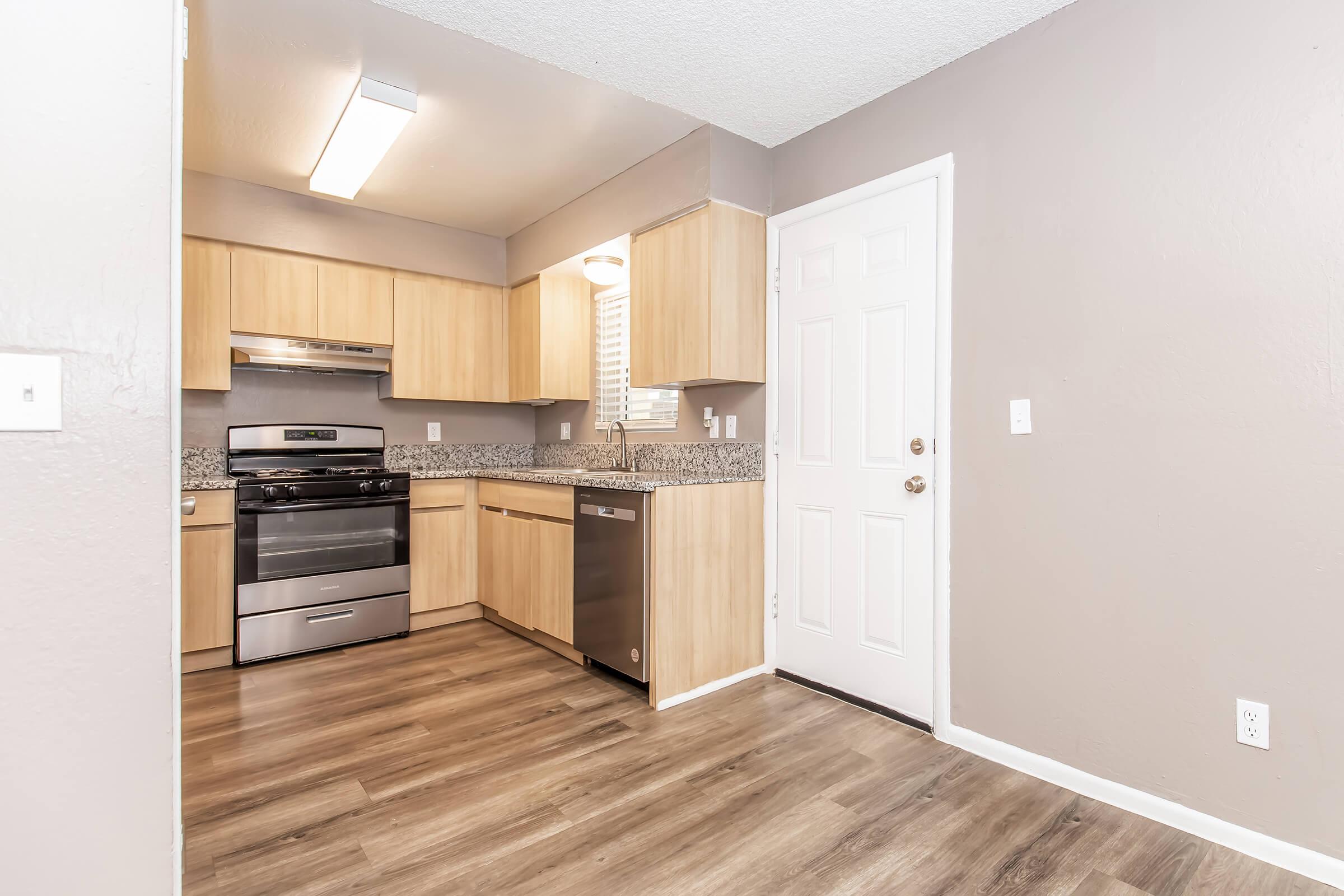 Modern kitchen with light wood cabinetry, granite countertops, stainless steel appliances including an oven, and a dishwasher. The room features a window for natural light, vinyl flooring, and a white door leading to an entryway.