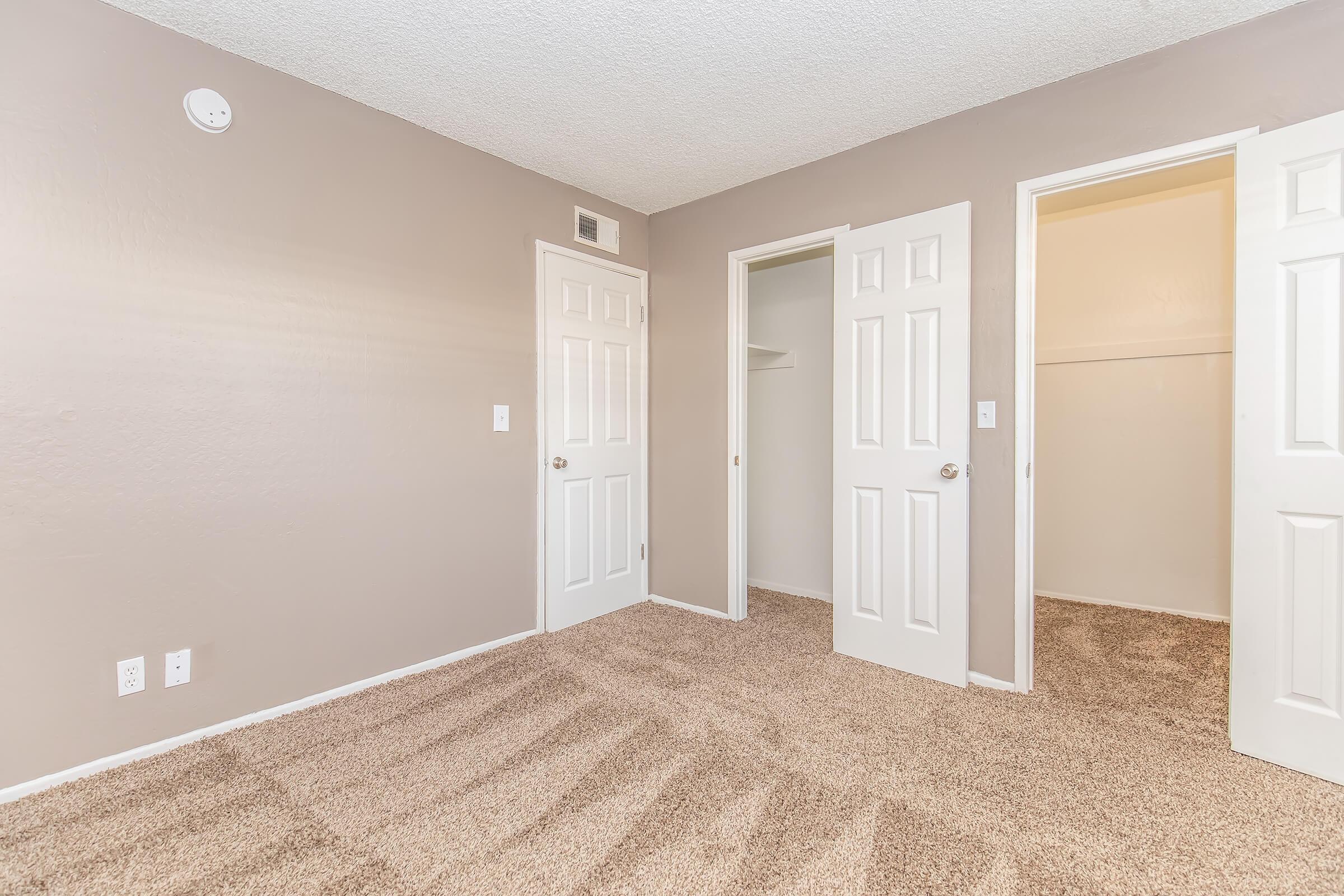 A spacious, empty bedroom with light beige walls and plush carpet. Three white paneled doors are visible: one is closed, one opens to a small closet, and the third opens to another room. The ceiling is textured, and there's a small white vent on one wall. Natural light illuminates the space.