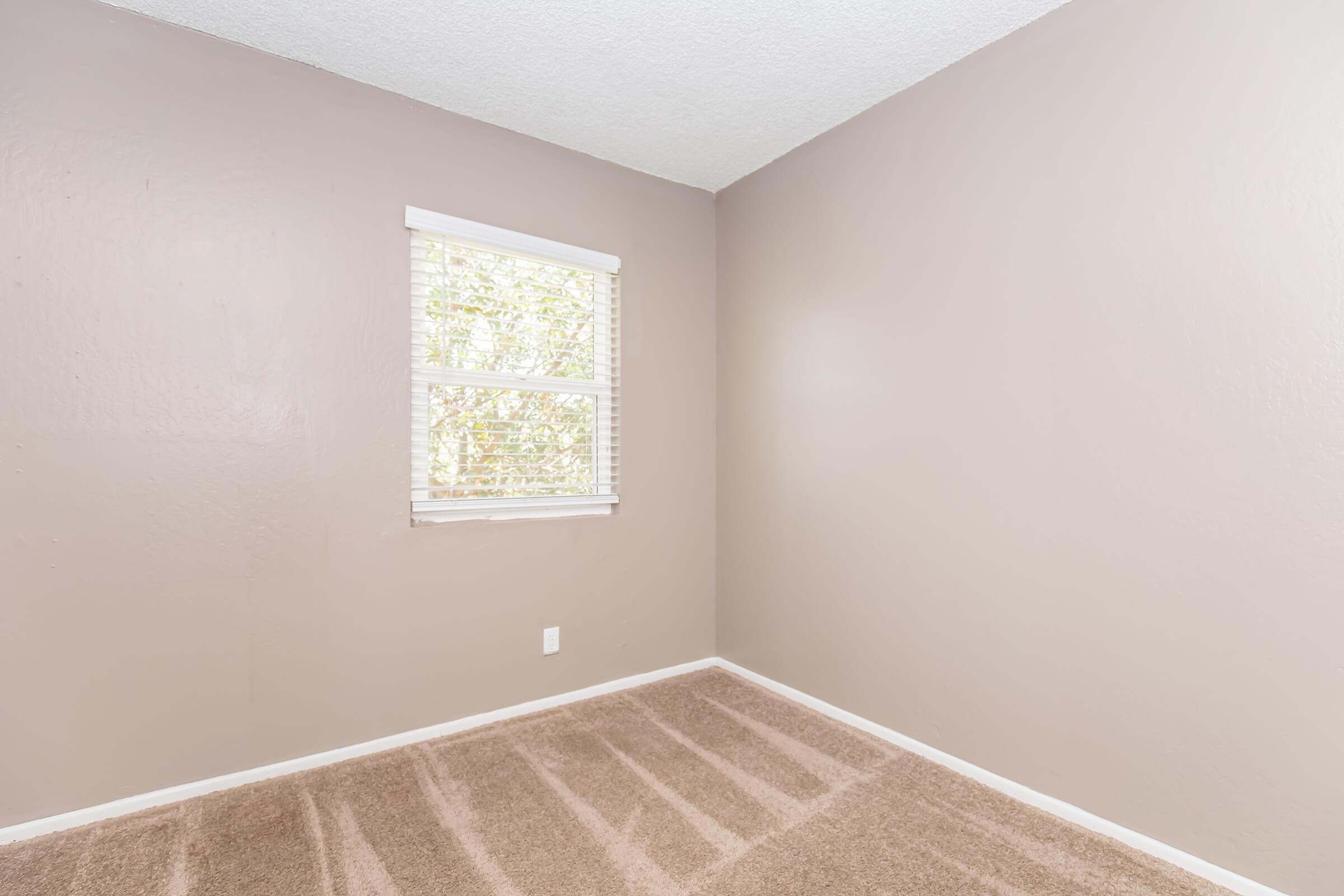 A small, empty room featuring light brown walls and a window with white blinds. The floor is covered in beige carpet with subtle patterns. The room is well-lit by natural light coming through the window, creating a simple and clean atmosphere.