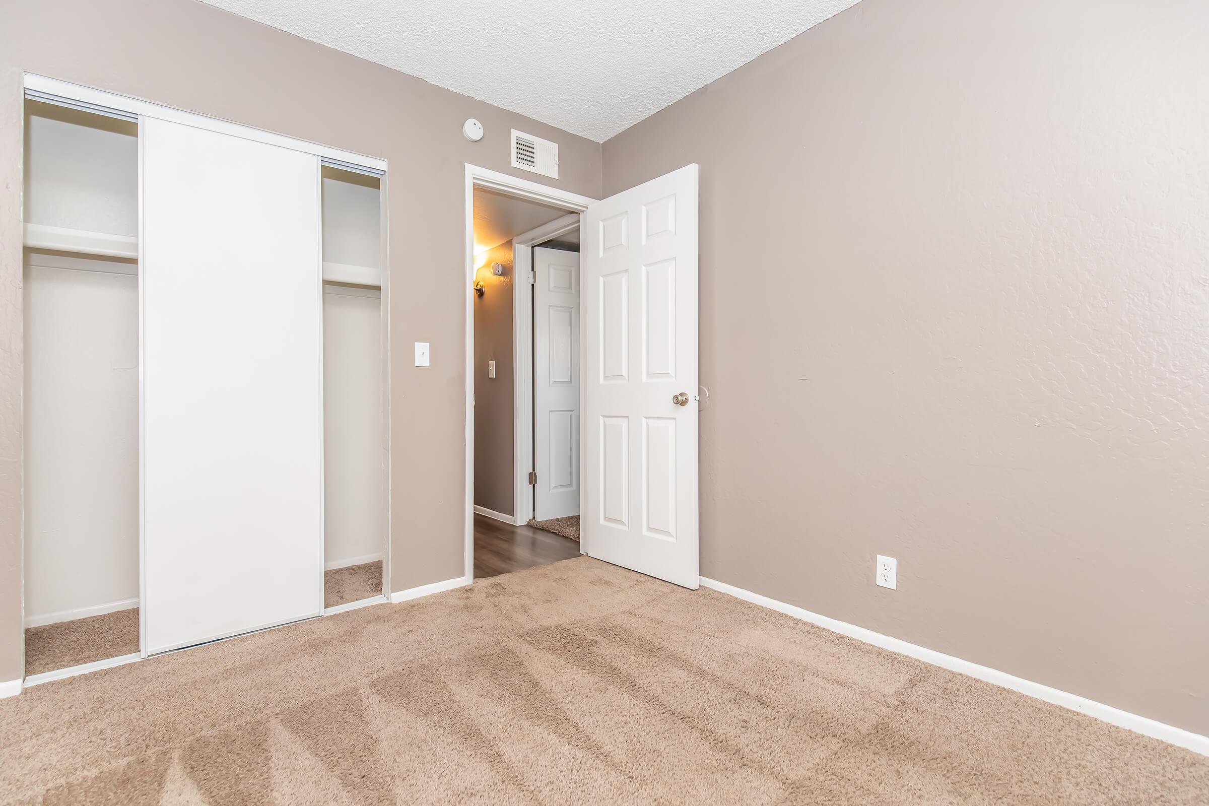 A well-lit room featuring beige walls and carpeted flooring. There is a sliding mirrored closet door on the left and a white door leading to another room on the right. The overall ambiance is neutral and inviting, suitable for a bedroom or study.