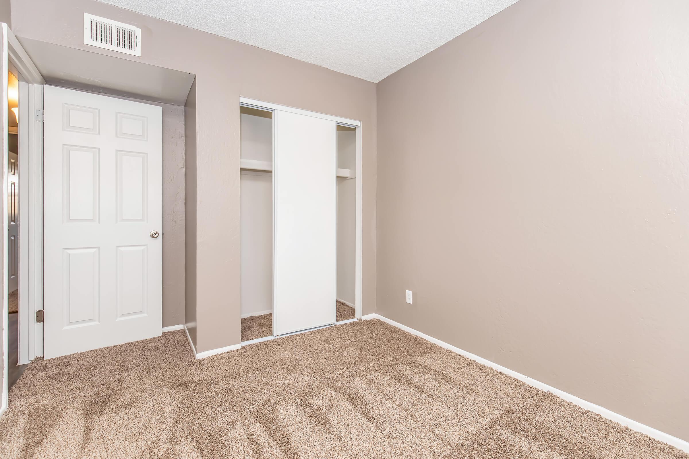 A simple, empty room with light brown carpet and beige walls. There is a white sliding door closet on the right and a closed white door on the left. The room is well-lit, with natural light coming from an unseen source, creating a clean and spacious feel.