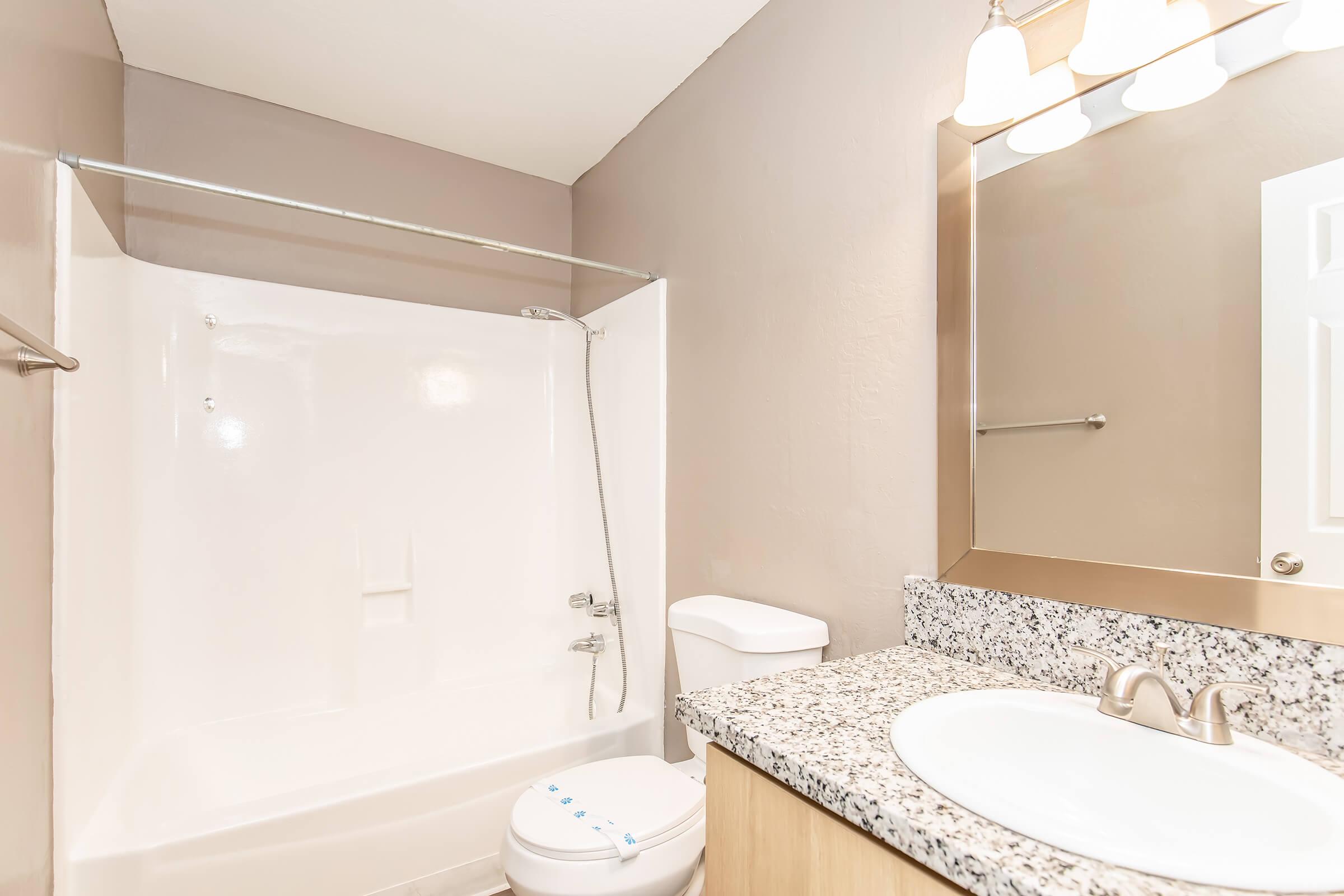 A clean bathroom featuring a white bathtub and shower, a modern vanity with a granite countertop, a white toilet, and a large mirror with three light fixtures above. The walls are painted in a neutral color, giving the space a bright and airy feel.