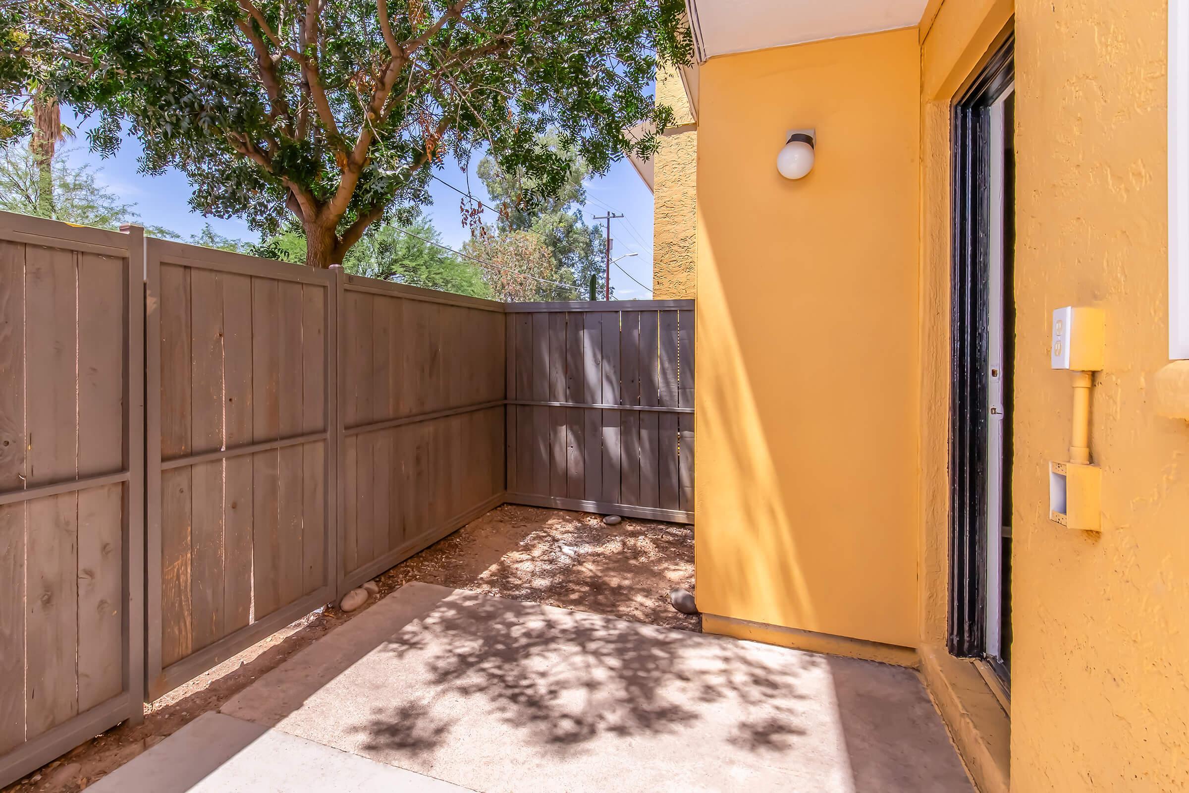 A corner patio area with a yellow wall and gray fencing. The ground is covered in gravel, and there is a tree casting shadows. A light fixture is mounted near the entrance door. The surrounding area appears well-maintained and secluded.