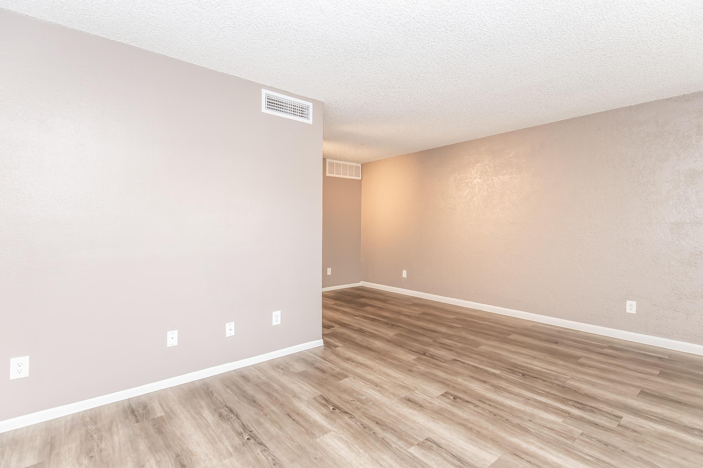 A spacious, empty room with light brown, textured walls and a light wooden floor. The room has no furniture, showcasing a clean and minimalist space. A vent is visible on one wall, and warm lighting illuminates the area, creating a welcoming atmosphere.