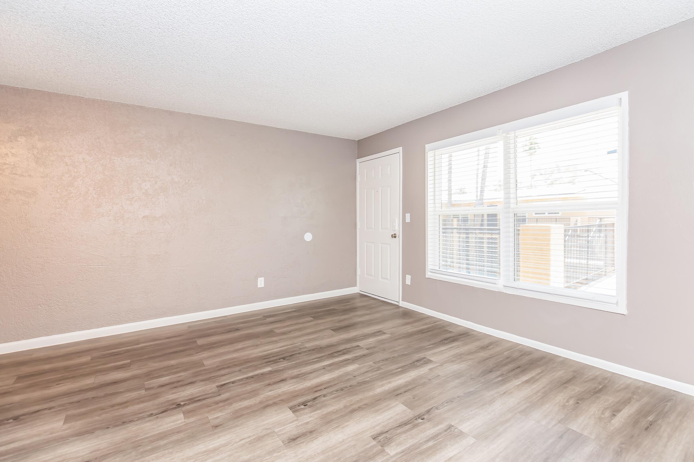 A spacious, empty room with light brown walls and new wooden flooring. There is a door on the right side leading outside and a large window with white blinds allowing natural light to enter. The overall ambiance is bright and neutral, suitable for various decorating styles.