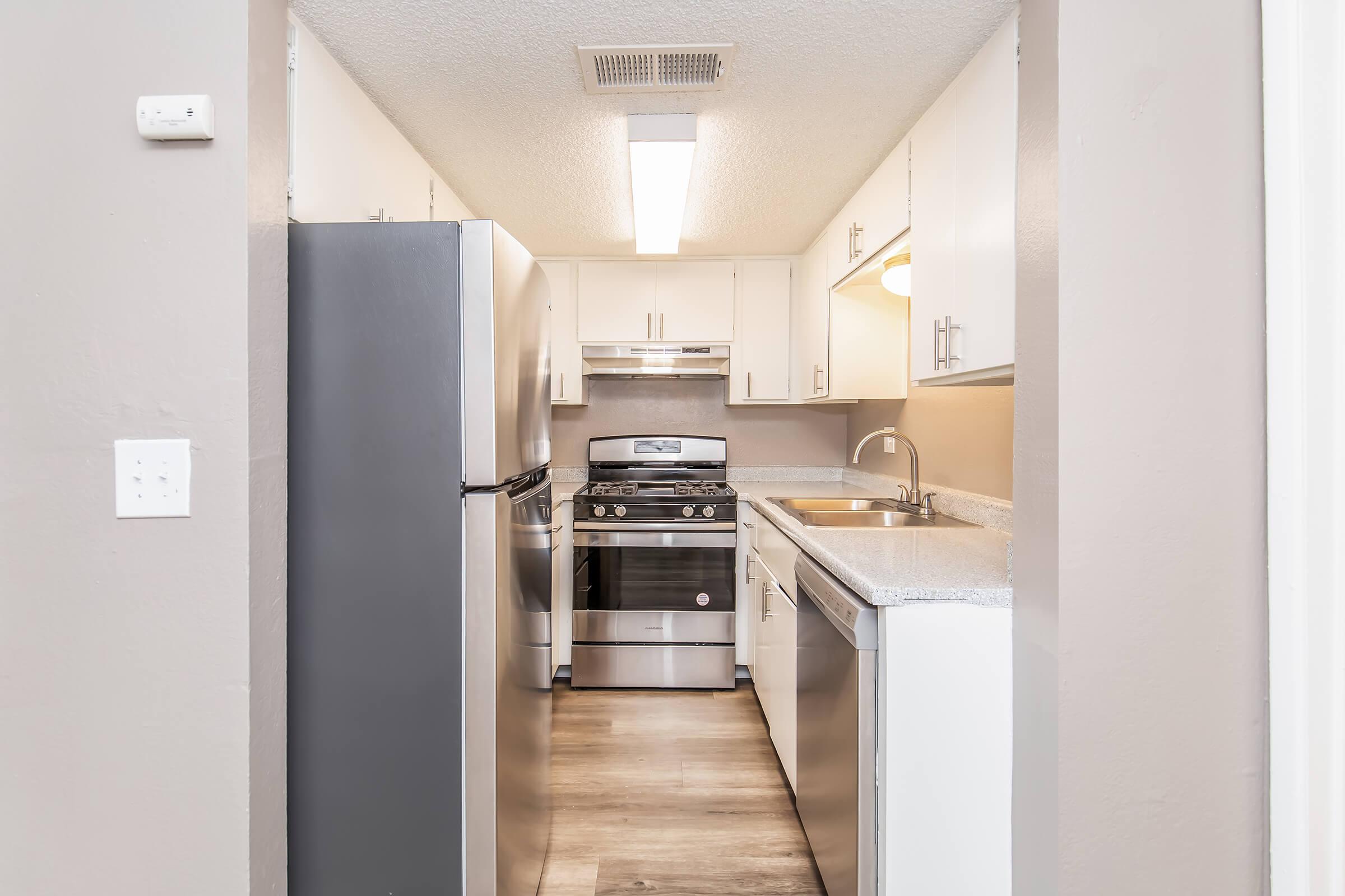 A modern kitchen viewed from a hallway, featuring stainless steel appliances including a refrigerator, stove, and dishwasher. The cabinets are white with a light countertop, and there is overhead lighting illuminating the space. The floor is a light wood finish, creating a clean and inviting atmosphere.
