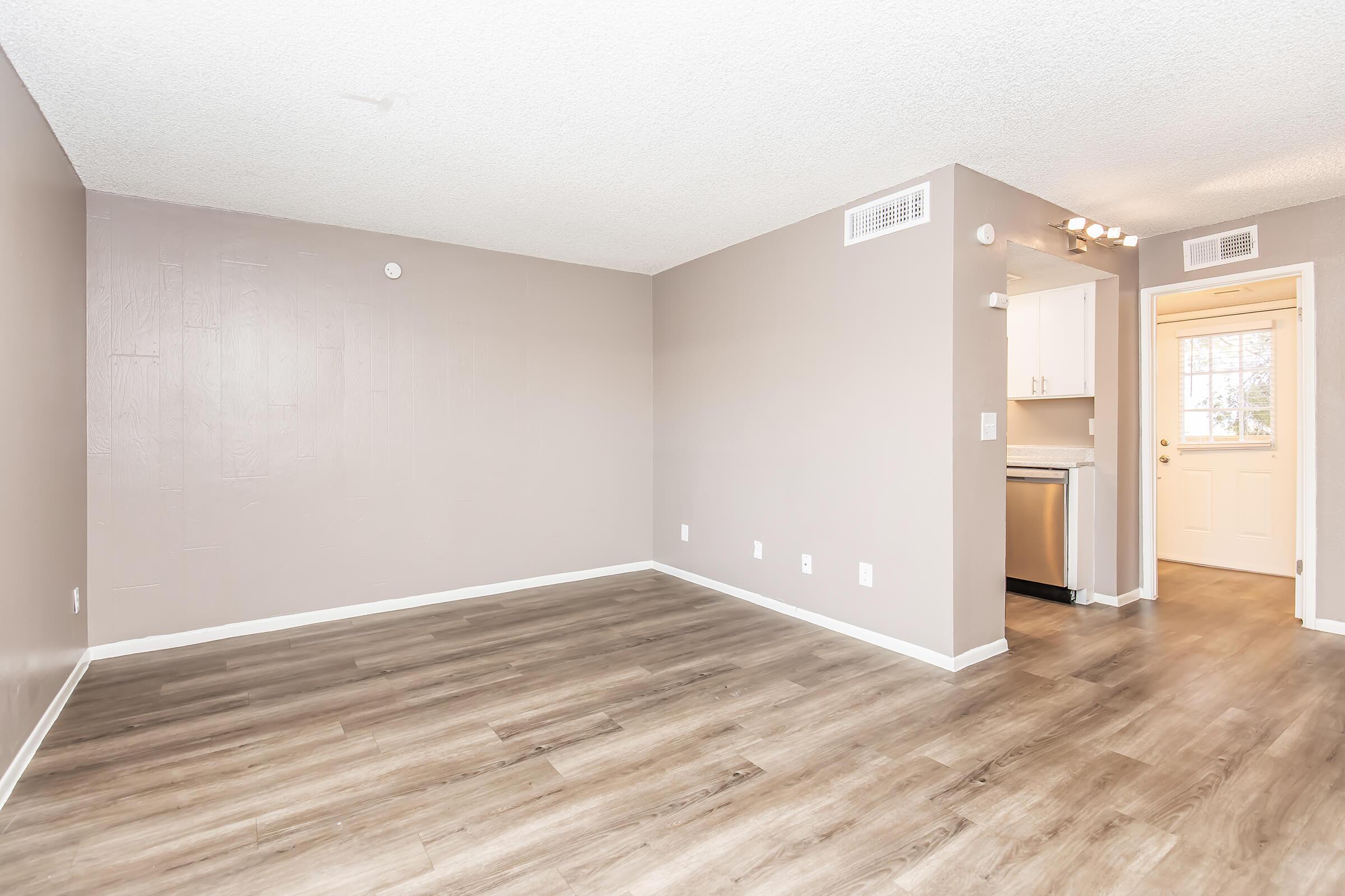Spacious living area with light brown laminate flooring, neutral-colored walls, and a doorway leading to a kitchen with modern appliances. Natural light flows in from the door, creating a bright atmosphere.