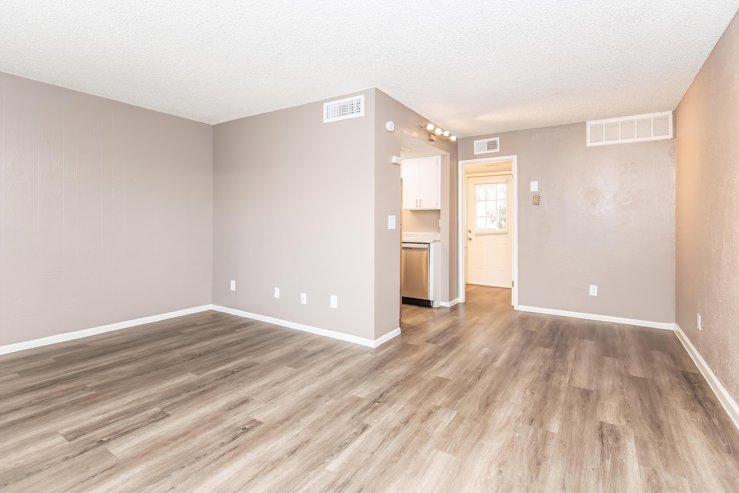 A spacious living area featuring light grey walls and new vinyl plank flooring. The room is empty, showcasing its ample space. To the right, there is an open doorway leading to a kitchen with white cabinetry. Natural light enters through the doorway, enhancing the room's bright and inviting atmosphere.