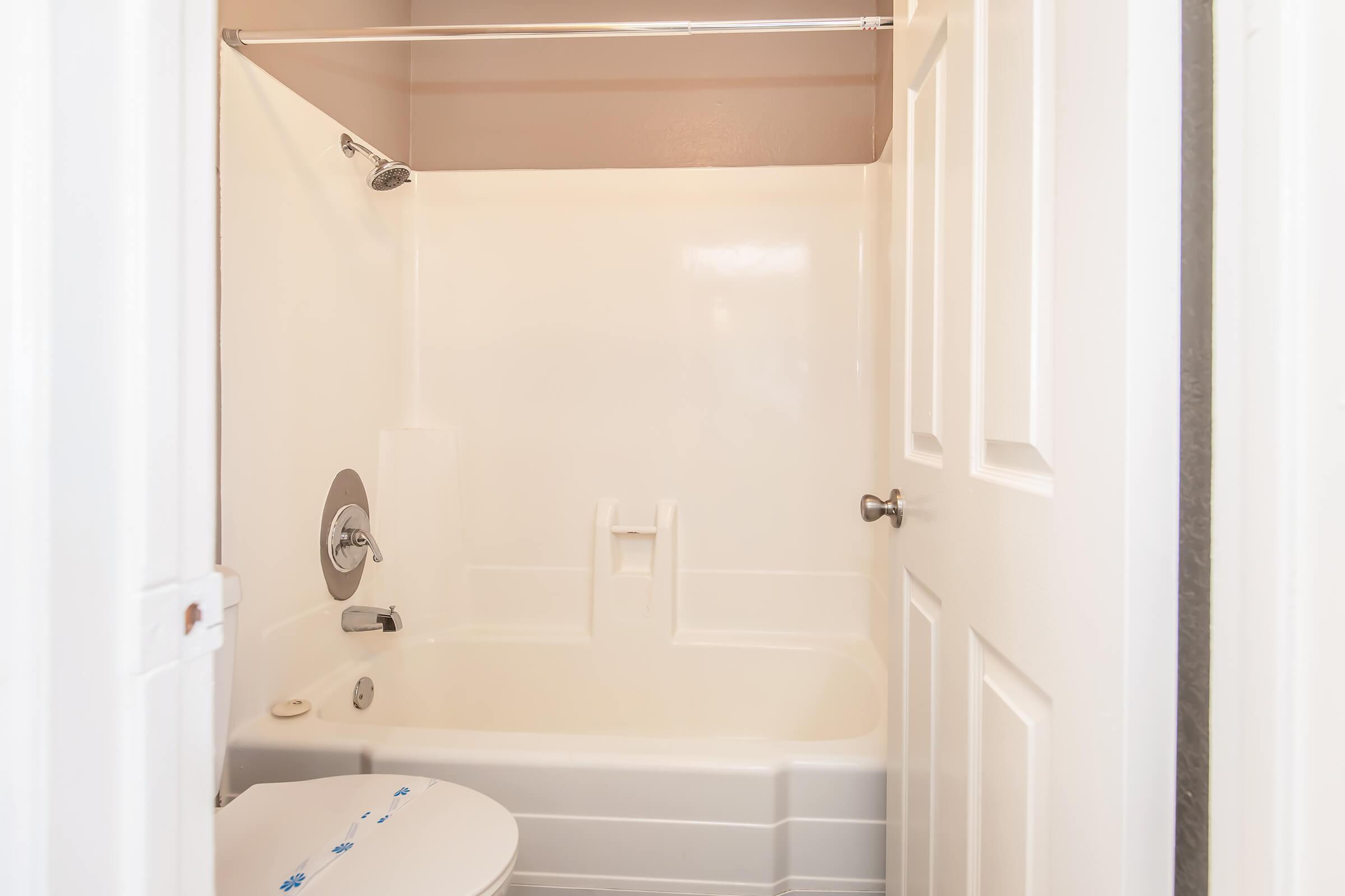 A small bathroom featuring a bathtub with a showerhead, a wall-mounted faucet, and a toilet. The walls are a light color, and there is a shower curtain rod above the bathtub. The space is clean and well-maintained, with white paneling and a door leading to another room.
