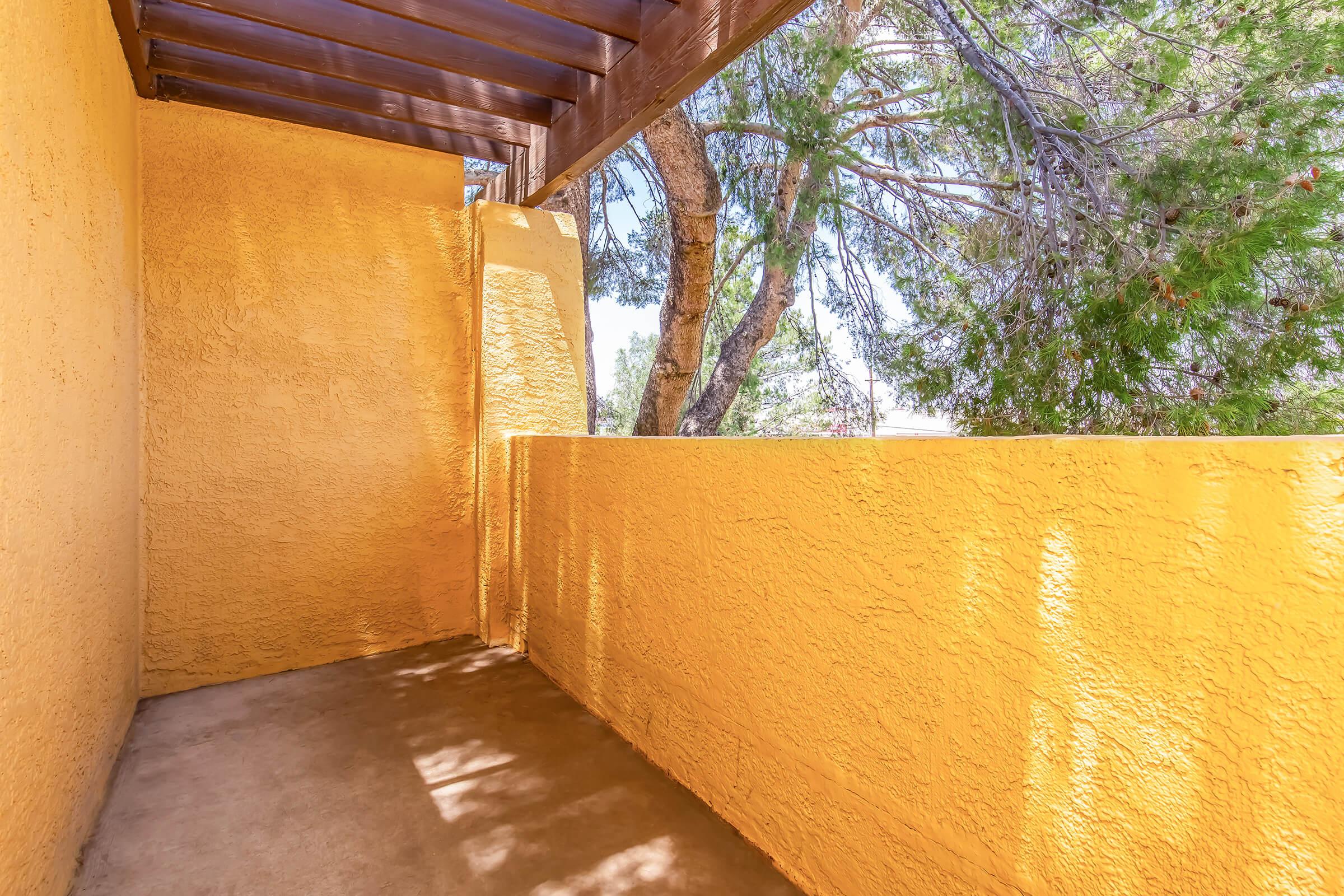 A sunlit balcony with textured yellow walls, surrounded by greenery from trees. The space is partly shaded, creating a warm and inviting atmosphere, with soft shadows playing on the floor.
