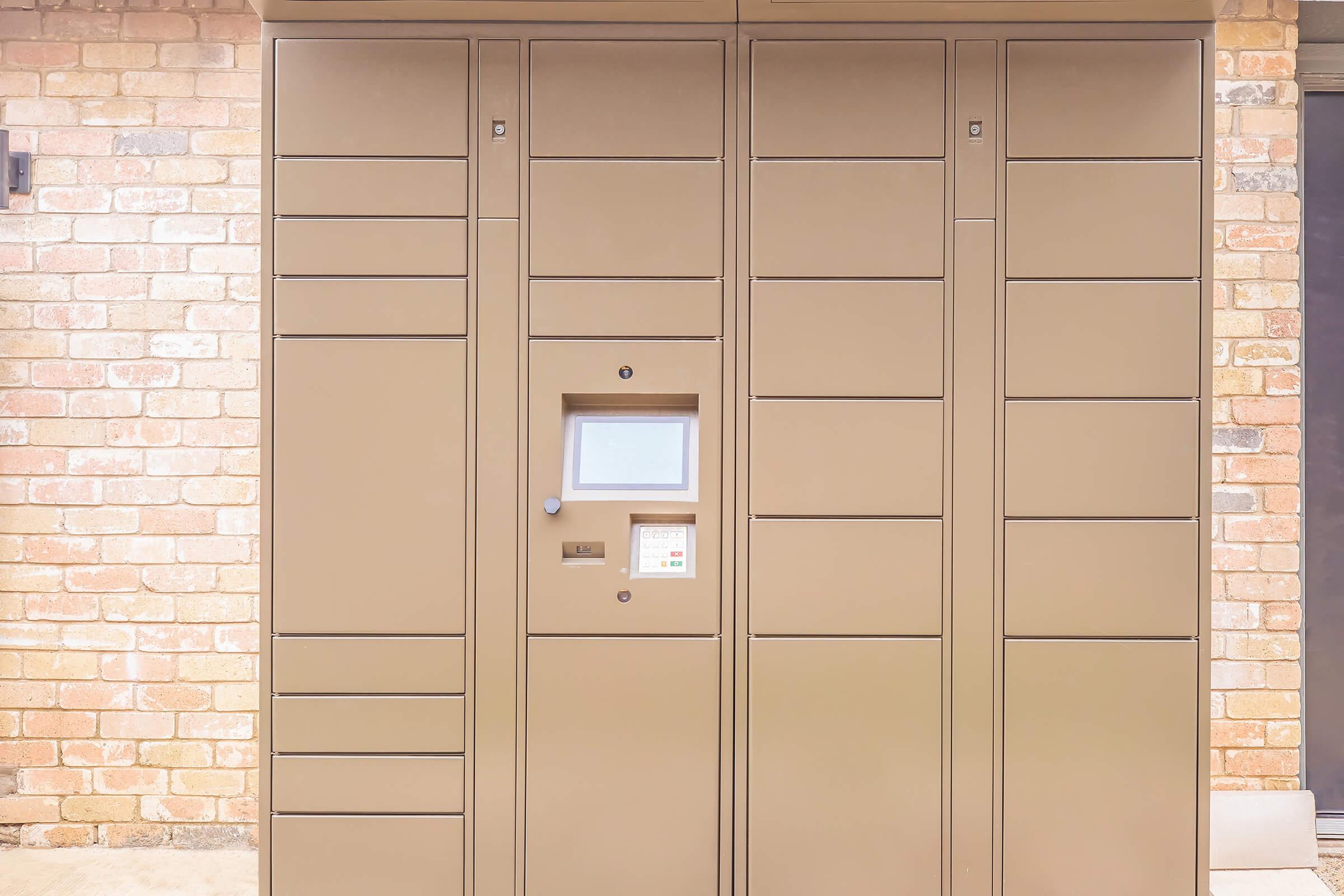A tan delivery locker unit against a brick wall, featuring a digital screen and buttons for package access. The locker is designed for secure package retrieval, with multiple compartments for different sizes of deliveries.