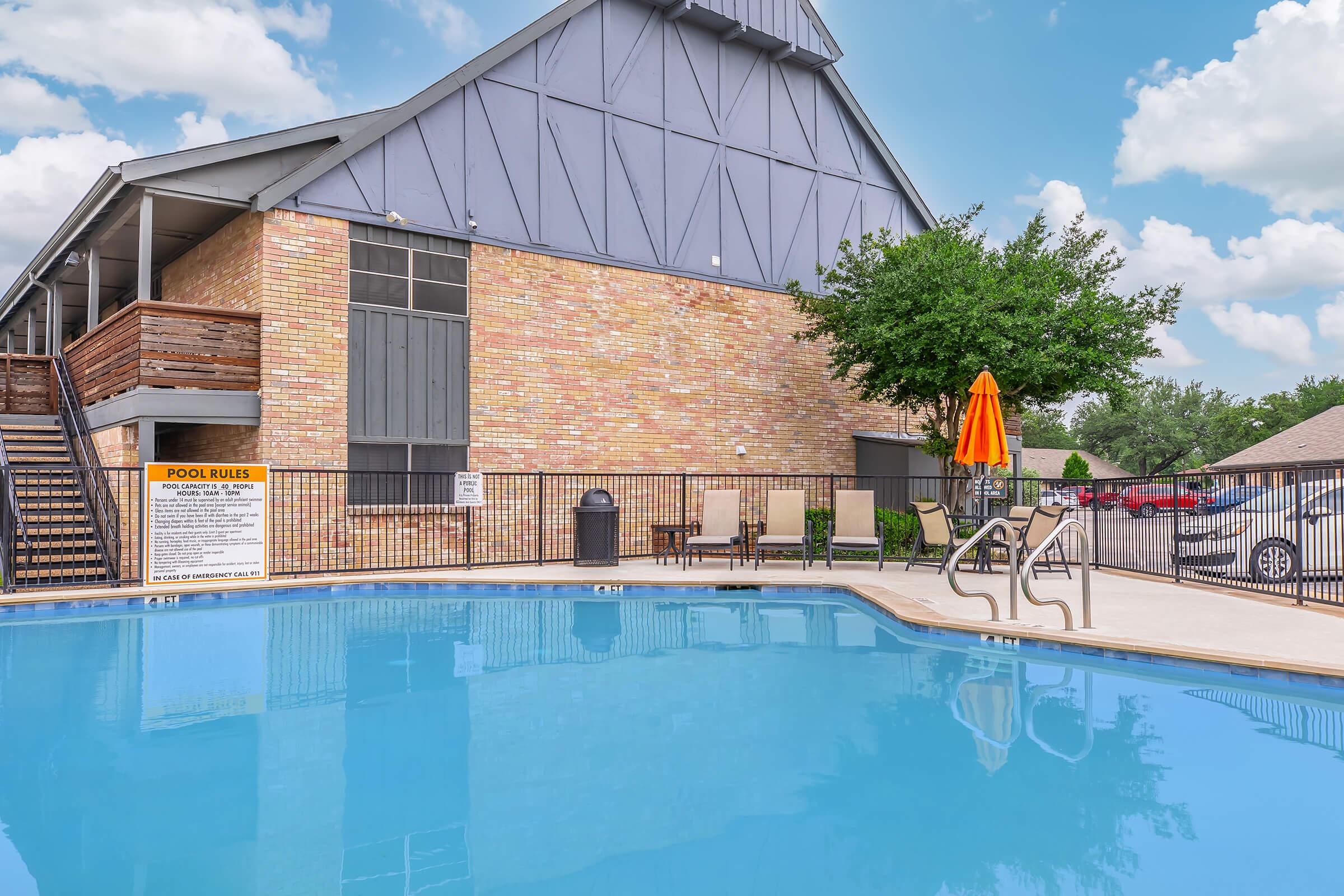 A clear blue swimming pool with a ladder in the foreground, surrounded by a black metal fence. A building with a brick facade and large windows is in the background, along with a green tree. An orange umbrella shades a seating area nearby, and pool rules are posted on a sign. The sky is partly cloudy.
