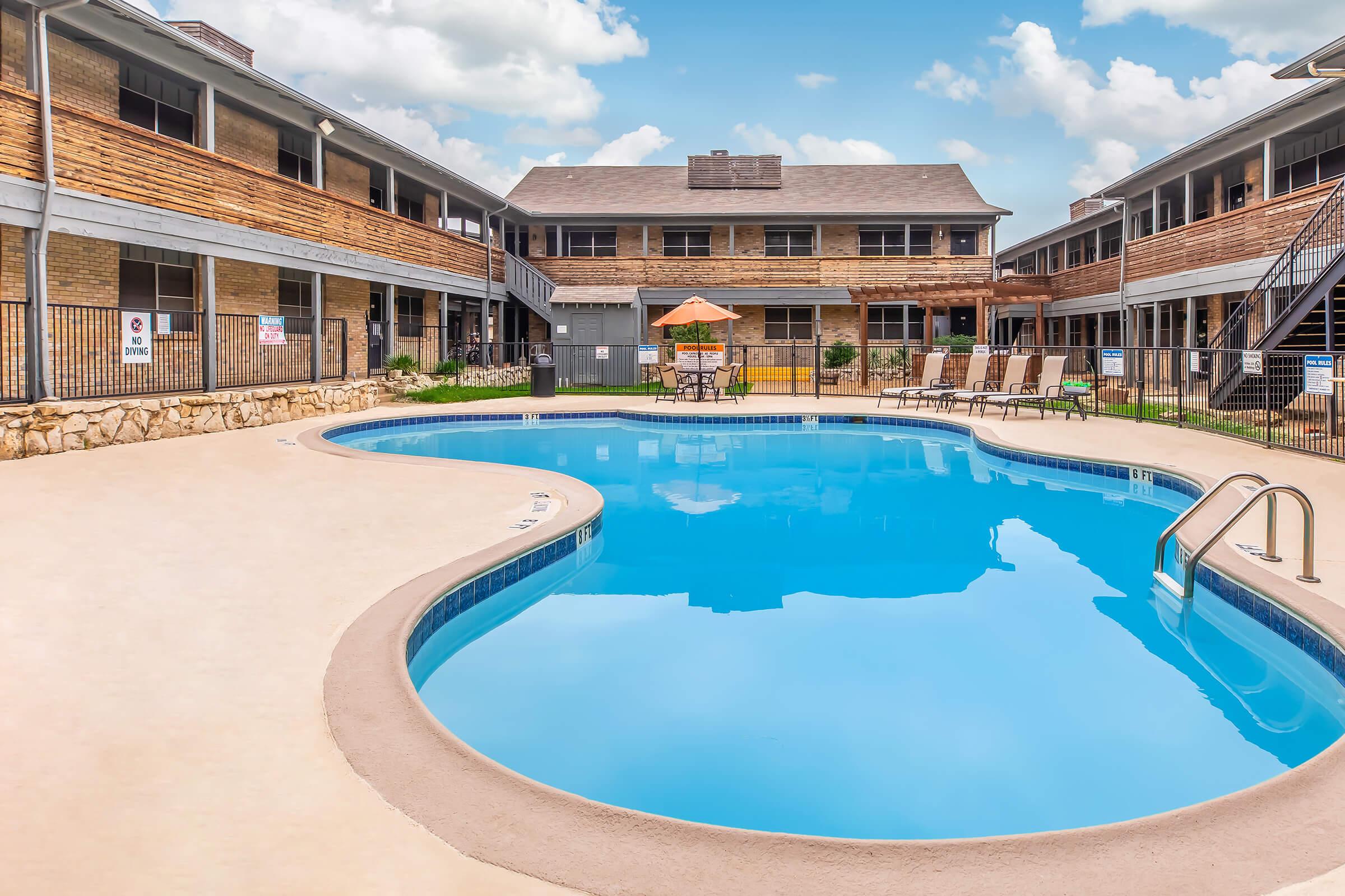 A clear blue swimming pool surrounded by sun loungers and an umbrella, with two-story buildings in the background. The area is well-maintained with green grass and a stone border along the pool. The sky is partly cloudy.