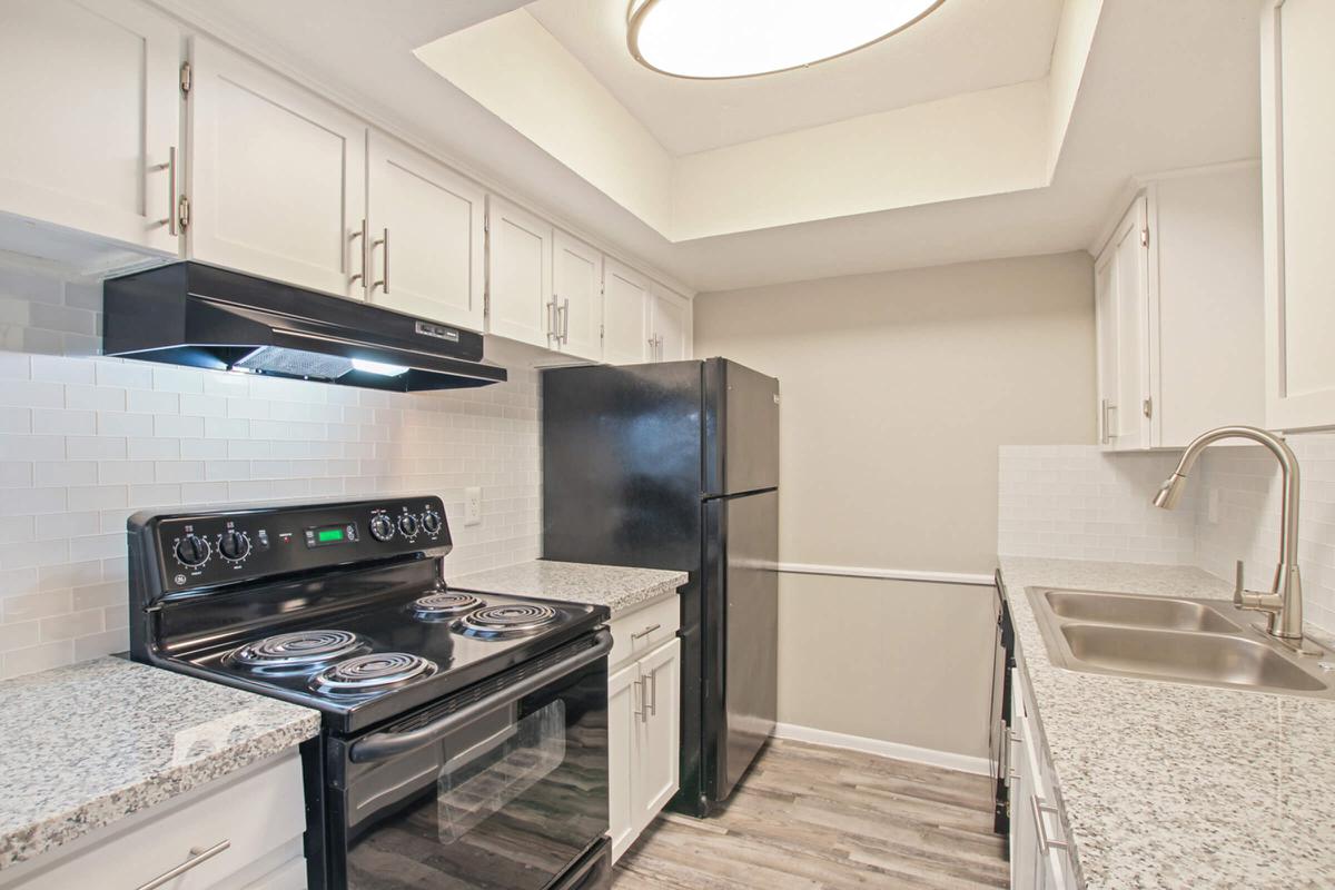 A modern kitchen featuring white cabinetry, a black refrigerator, and an oven with a hood above it. The countertops are made of speckled grey granite, and there is a stainless steel sink under a window. The ceiling has a circular light fixture, and the backsplash is made of white tiles.