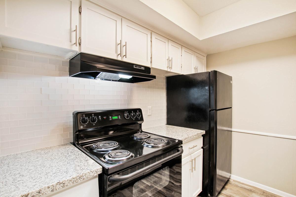 A modern kitchen featuring a black stove with four burners, an overhead microwave, and a black refrigerator. The cabinetry is white with silver handles, and the countertops are gray granite. There is a light-colored backsplash made of small tiles, creating a clean and contemporary look.