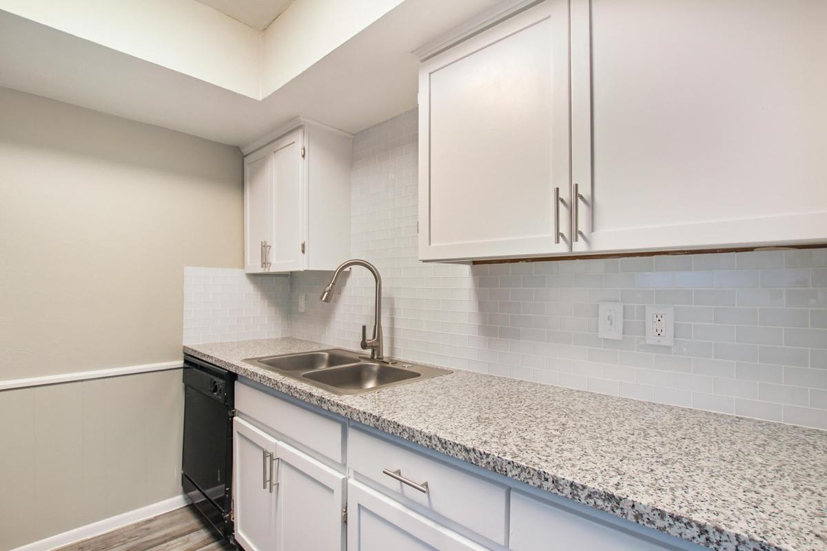 A modern kitchen featuring white cabinets, a gray granite countertop, and a stainless steel double sink. The backsplash is adorned with white tiles, and there's a black dishwasher visible. The space is well-lit with a neutral color scheme, creating a clean and inviting atmosphere.