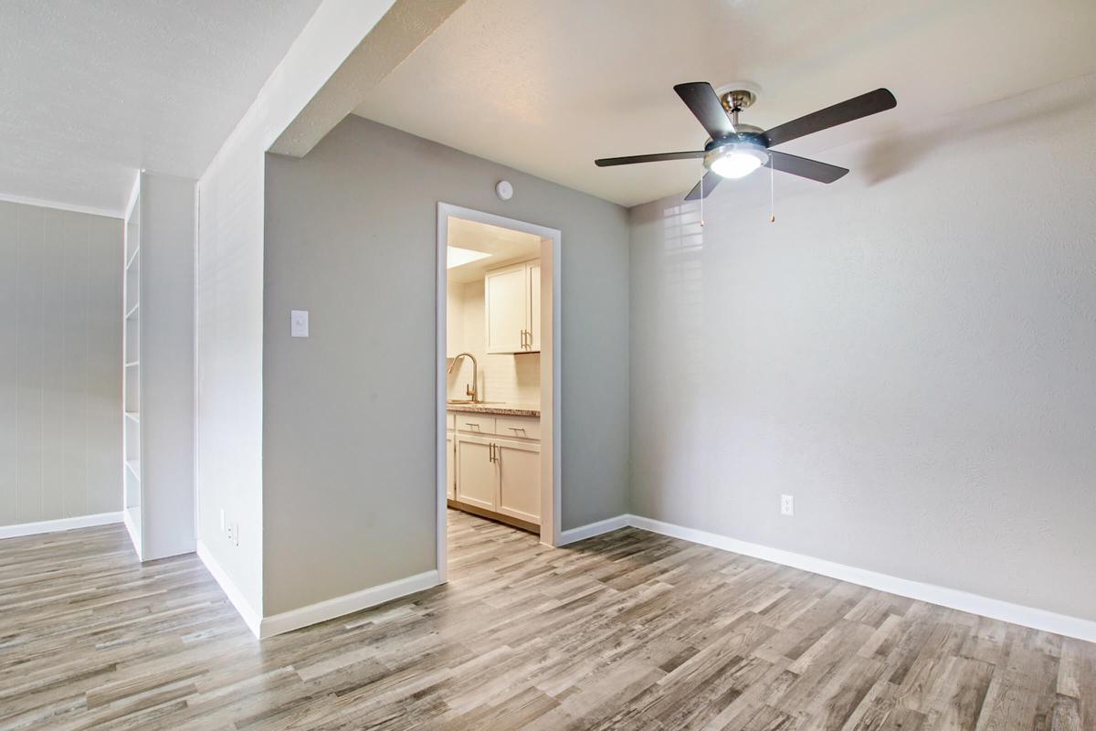 A bright, modern living space featuring light gray walls, a ceiling fan, and wooden-style flooring. An open doorway leads to a kitchen with white cabinets. The room is well-lit, creating a welcoming atmosphere. The design is minimalistic, ideal for comfortable living.