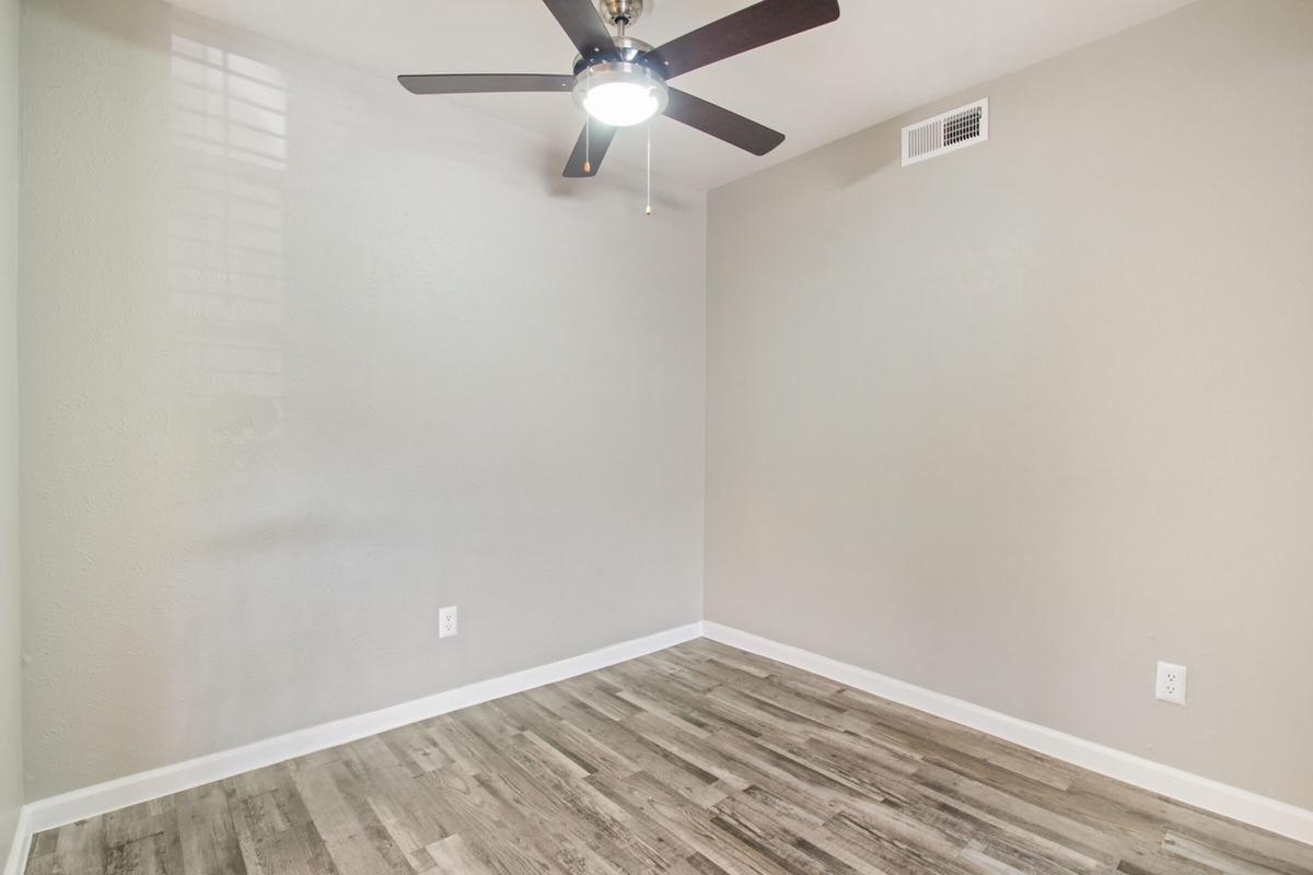 A vacant room with light gray walls and a ceiling fan. The floor features laminate planks in a natural wood finish. There are no furnishings or decorations, creating a spacious and minimalistic appearance. A window is present but not visible in the image, allowing for potential natural light.