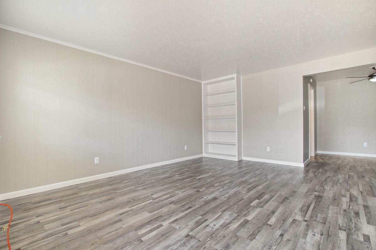 A spacious, empty living room featuring light-colored walls and a wood-like floor. One side has a built-in shelf, and there is a doorway leading to another room. Natural light illuminates the area, emphasizing the simplicity and openness of the space.