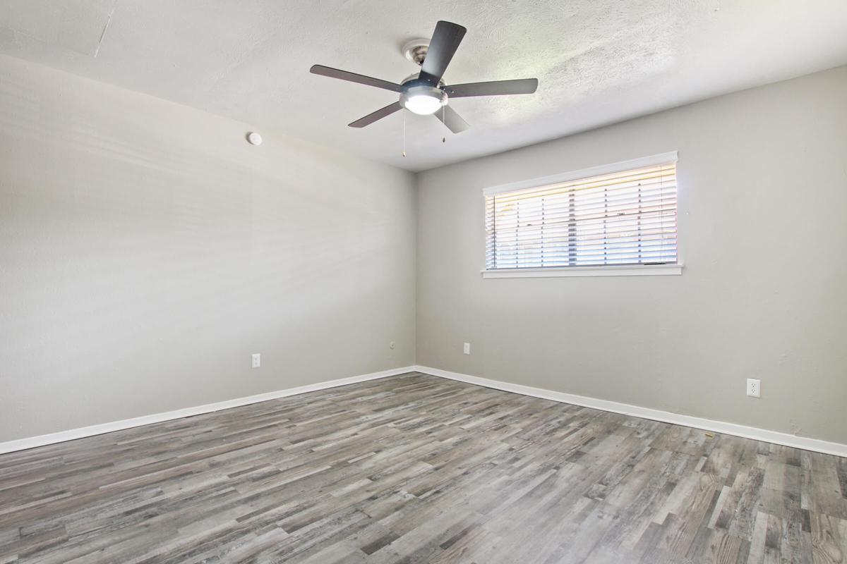 Empty room with gray walls and a ceiling fan. The floor is covered in light-colored wood laminate. A window with blinds allows natural light to enter the space, which has no furniture or decoration, creating a blank canvas for potential use.