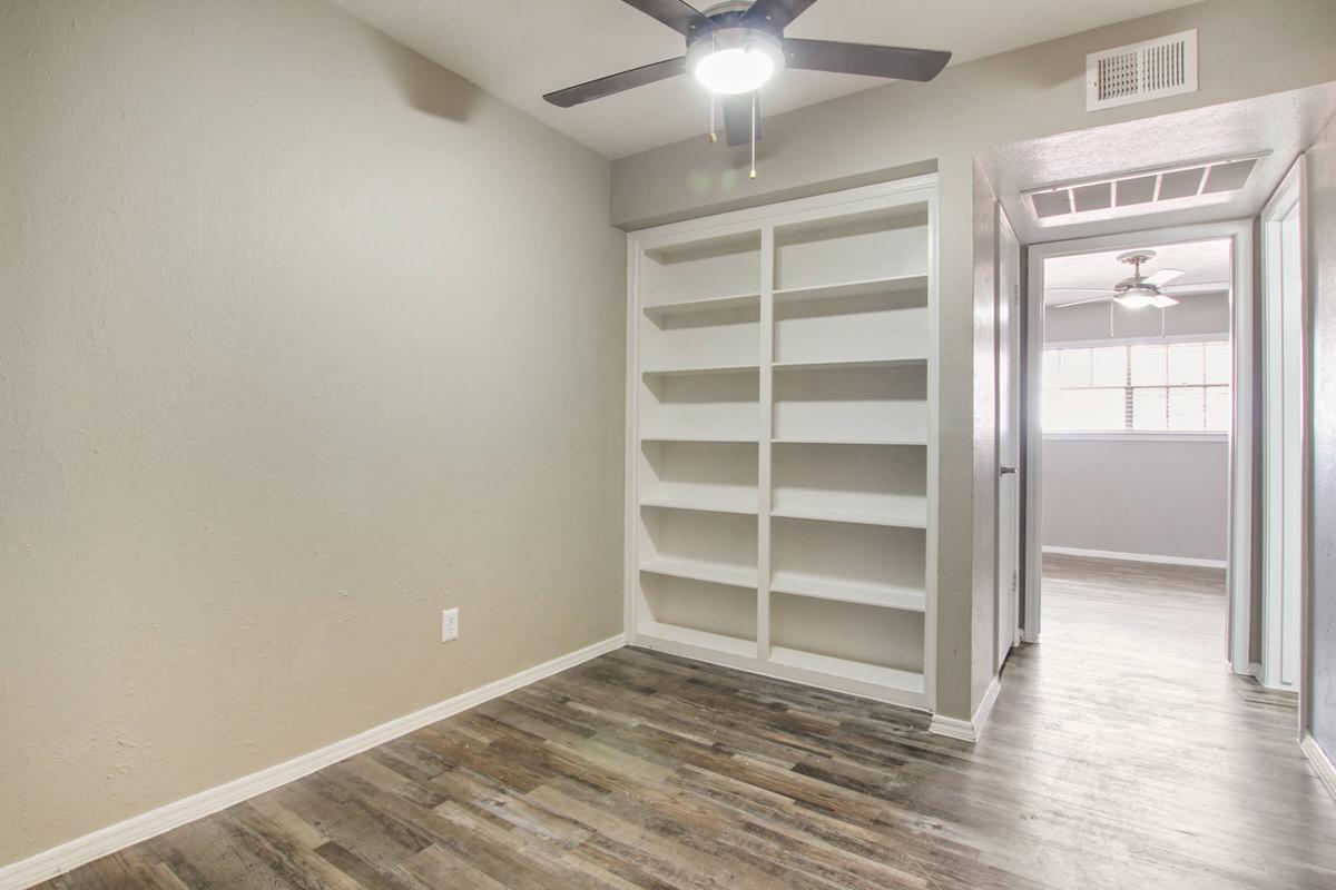 A vacant room with light gray walls and a ceiling fan. There is a built-in white shelf along one wall, and a doorway leading to another room. The flooring is a dark wood-like material, and there are windows allowing natural light to enter from the adjacent space.