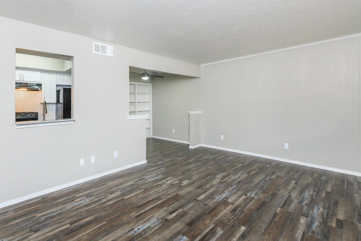 A spacious living area with light-colored walls and modern wood-like flooring. To the left, there is an open view into a kitchen with a bar area. The room features a ceiling fan and built-in shelves on the right. The overall ambiance is bright and inviting, suitable for various furniture arrangements.