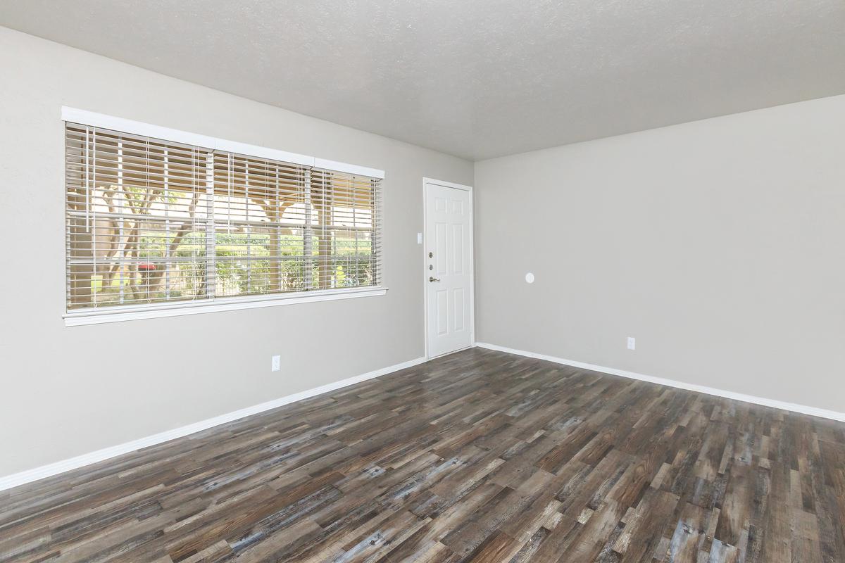 Interior of a room with light gray walls and a wooden floor. A large window with white blinds allows natural light to enter, and a white door is visible on the right. The space is empty and uncluttered, providing a clean and modern look.