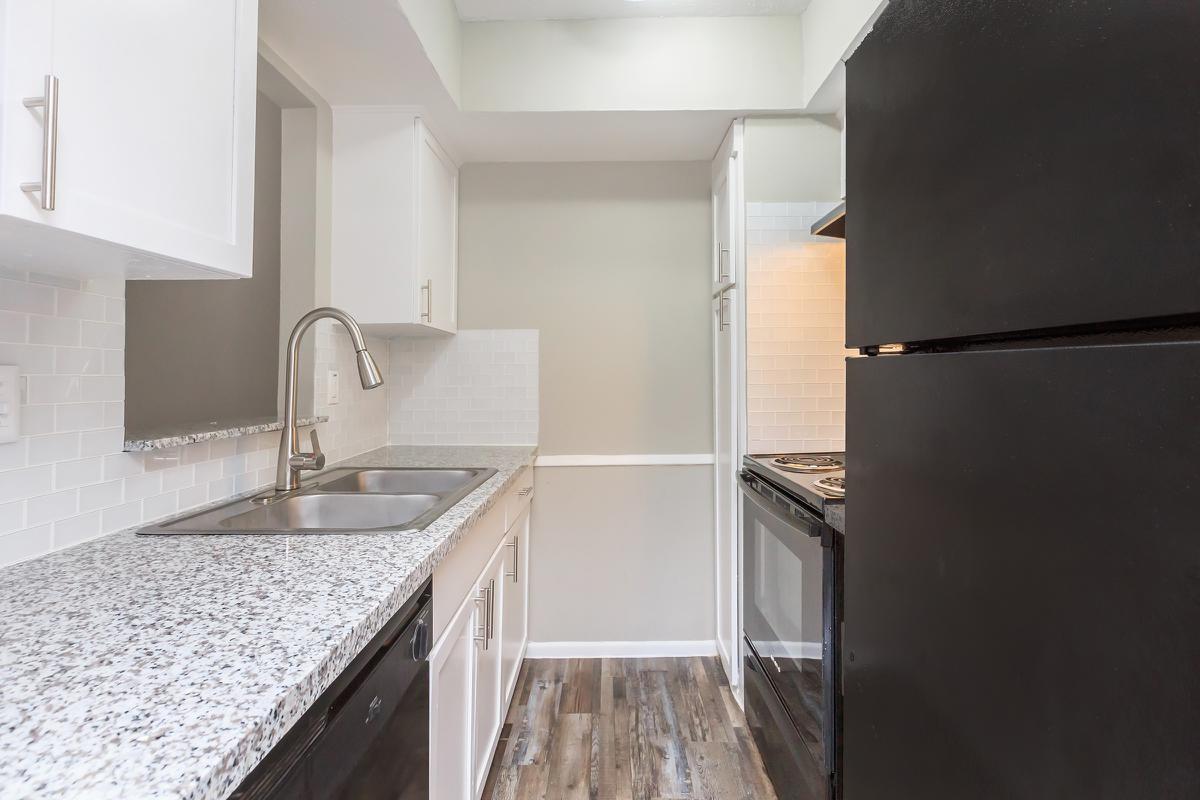 A modern kitchen featuring a sleek design with dark appliances, granite countertops, and white cabinetry. The space includes a double sink, a stovetop, an oven, and a refrigerator, complemented by a light-colored wall and wood-like flooring, creating a contemporary and inviting atmosphere.