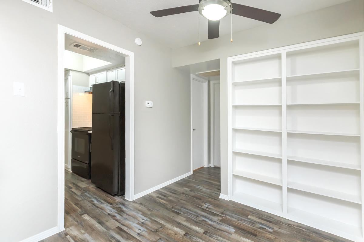 A view of a small room featuring a ceiling fan, light-colored walls, and a wooden floor. On one side, there is an open doorway leading to a kitchen with black appliances, and on the opposite side, built-in white shelves are mounted on the wall. The layout suggests it's a cozy living or office space.