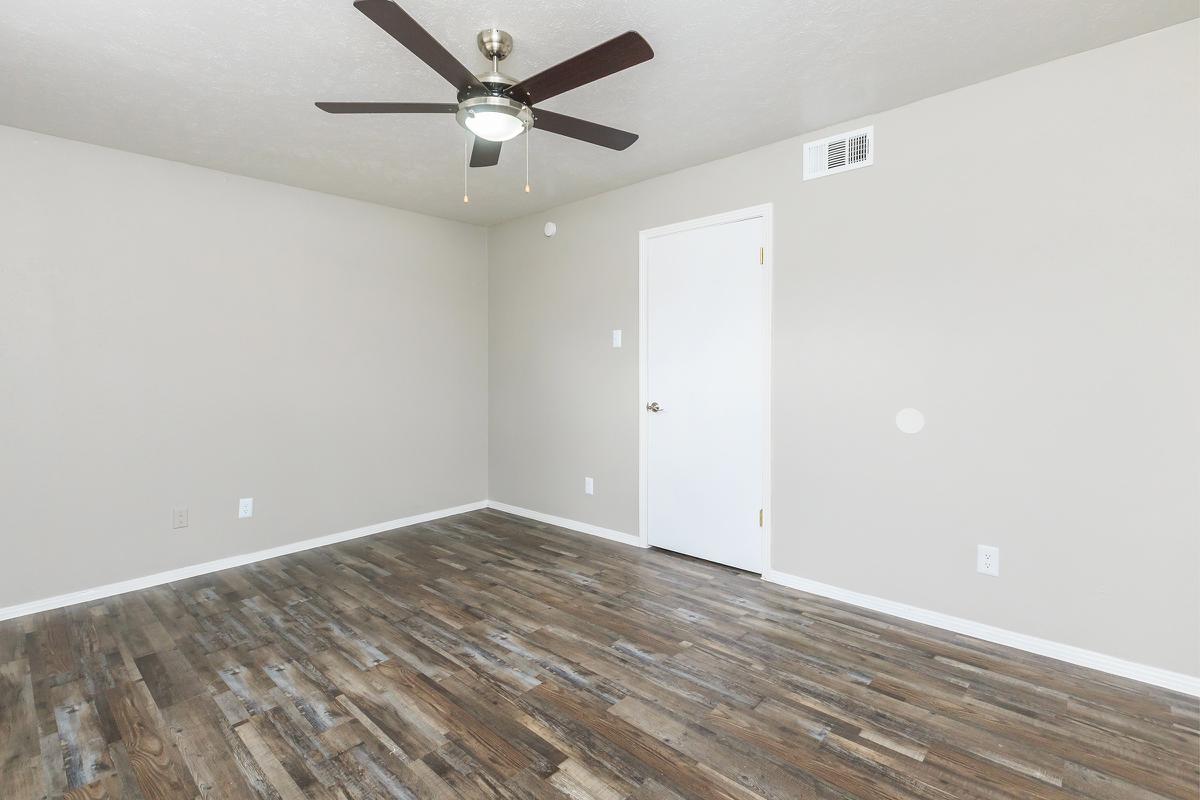 A spacious, empty room with light beige walls and a ceiling fan. The floor features wood-like laminate planks. A single white door is visible on the right side, and the room is well-lit with natural light. There are no furnishings or decorations in the space, creating a blank canvas for customization.