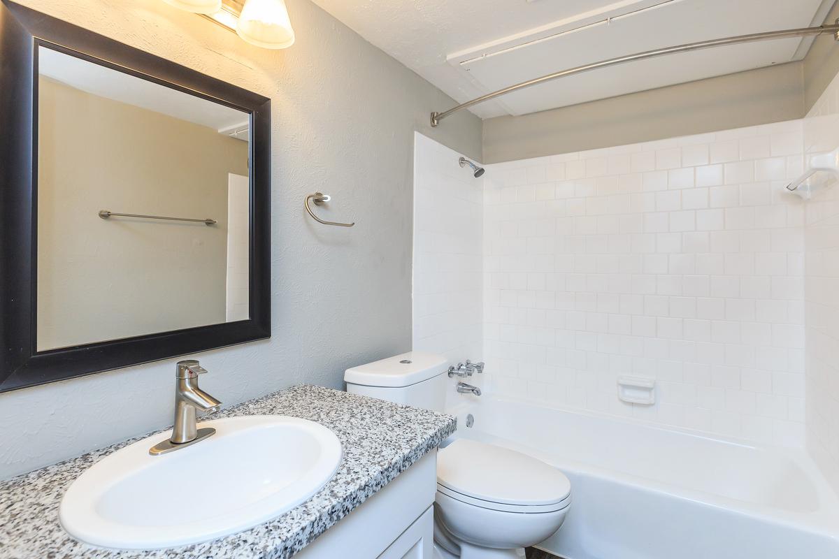 A modern bathroom featuring a white tiled bathtub with a shower curtain, a granite countertop sink with a sleek faucet, a mirror above the sink, and a toilet. The walls are painted in a neutral color, and there is a towel bar mounted beside the mirror. Soft lighting highlights the space.