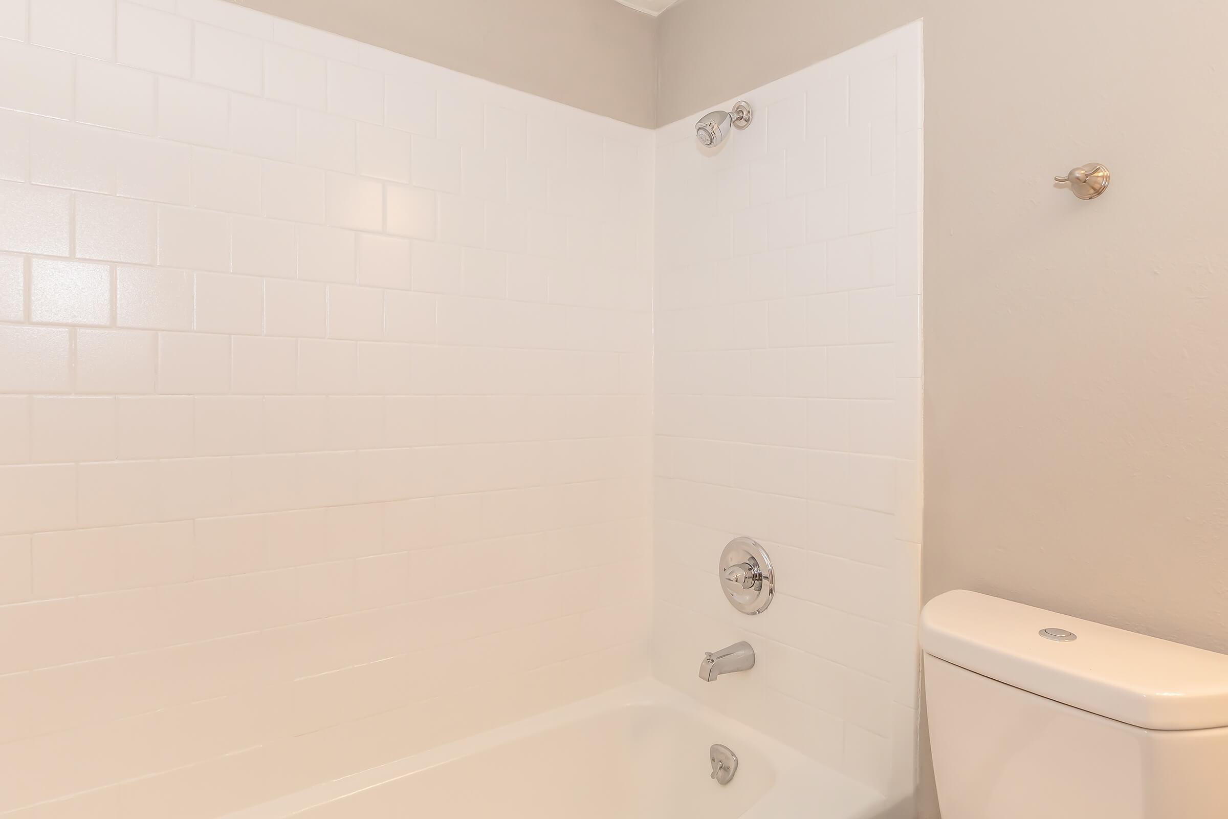 A clean, modern bathroom featuring a white tiled bathtub with a shower head, a chrome faucet, and a white toilet. The walls are a light gray color, and there is a simple design with no clutter, creating a spacious and inviting atmosphere.