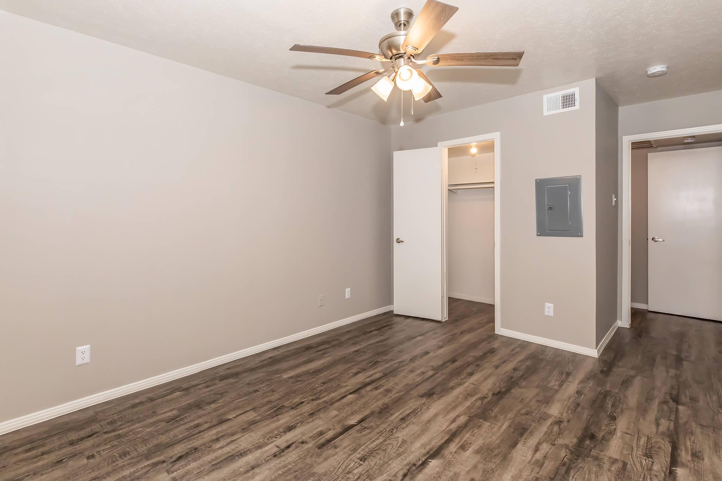 Empty room featuring light brown walls and flooring, a ceiling fan with light fixtures, a small closet with white doors, and a bathroom door on the right. The room has a neutral color palette and ample space, suitable for various furniture arrangements.
