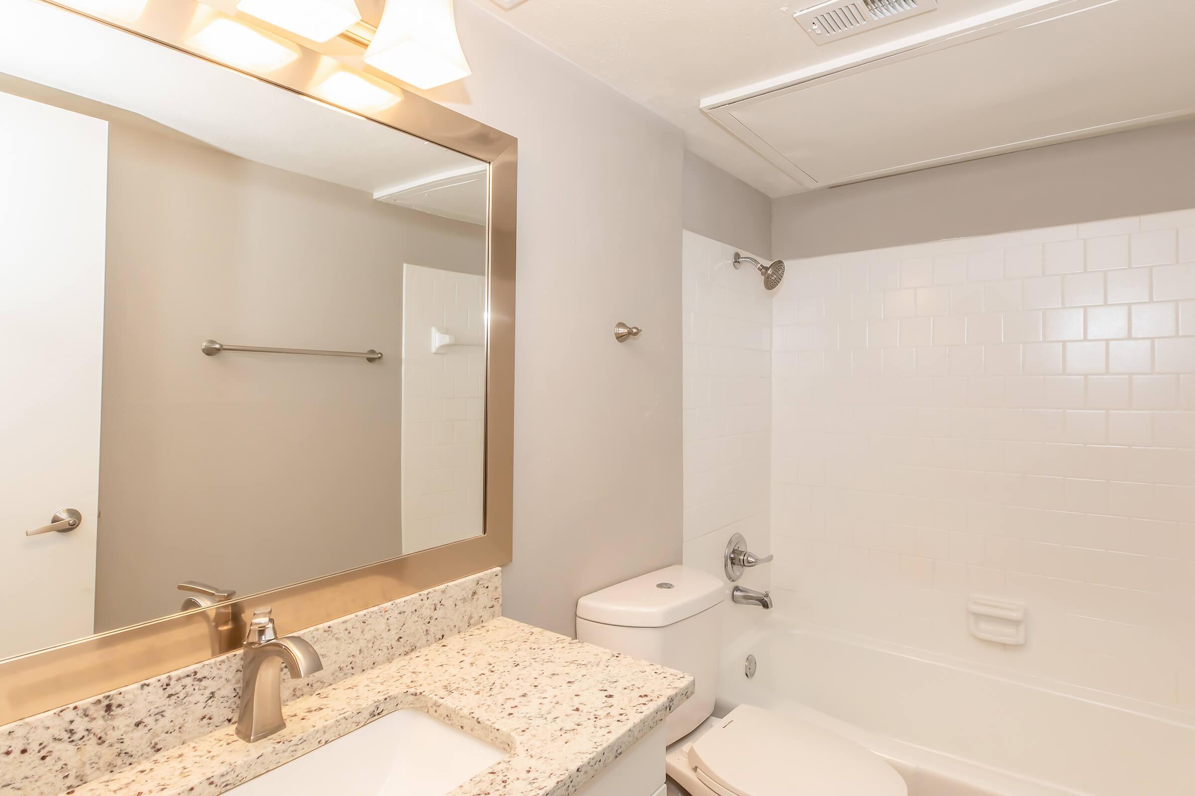 A clean and modern bathroom featuring a bathtub with a white tiled surround, a granite countertop with a sink, a large mirror above the sink, and a towel rack on the wall. The walls are painted in a light gray color, and there is natural light coming from an adjacent room.