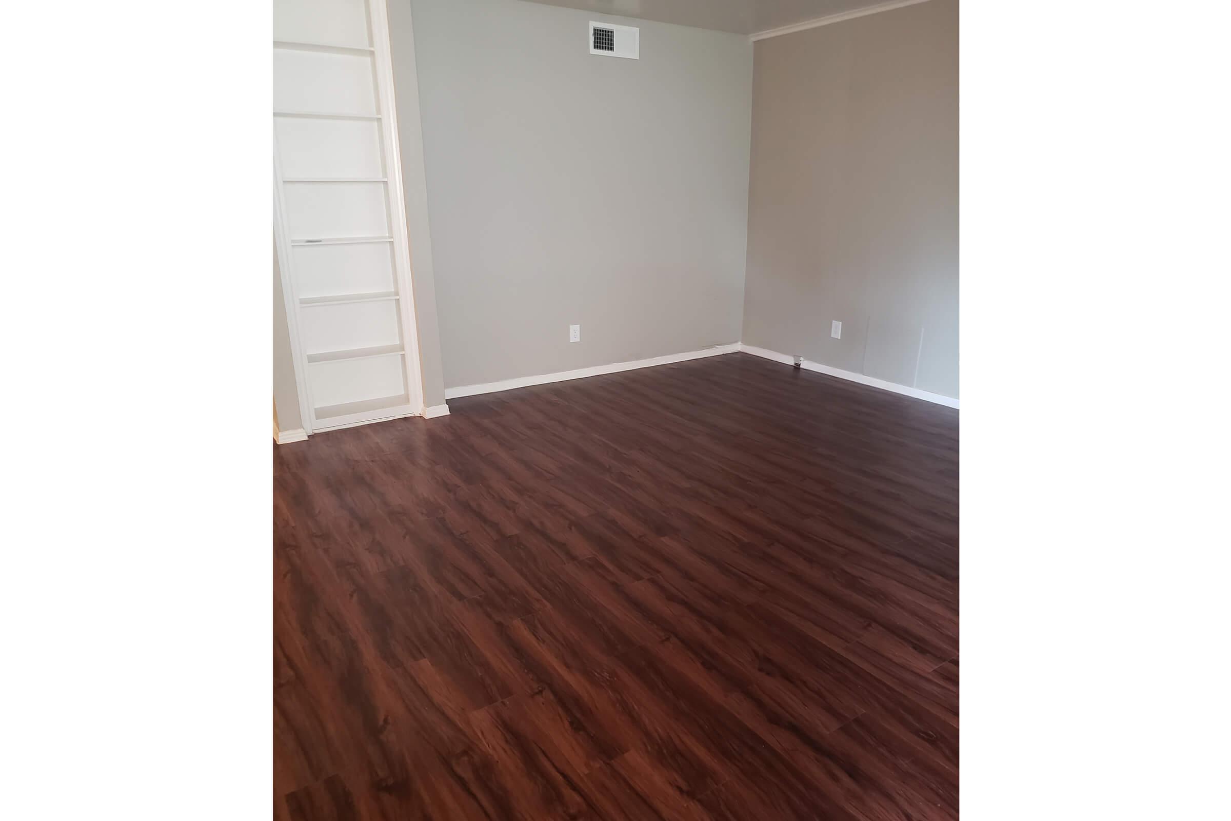 A spacious room featuring a smooth, dark wood laminate floor. The walls are painted light gray, and there is a small white built-in shelf or closet on the left side. The room appears bright and uncluttered, with a neutral color scheme that enhances the sense of openness.