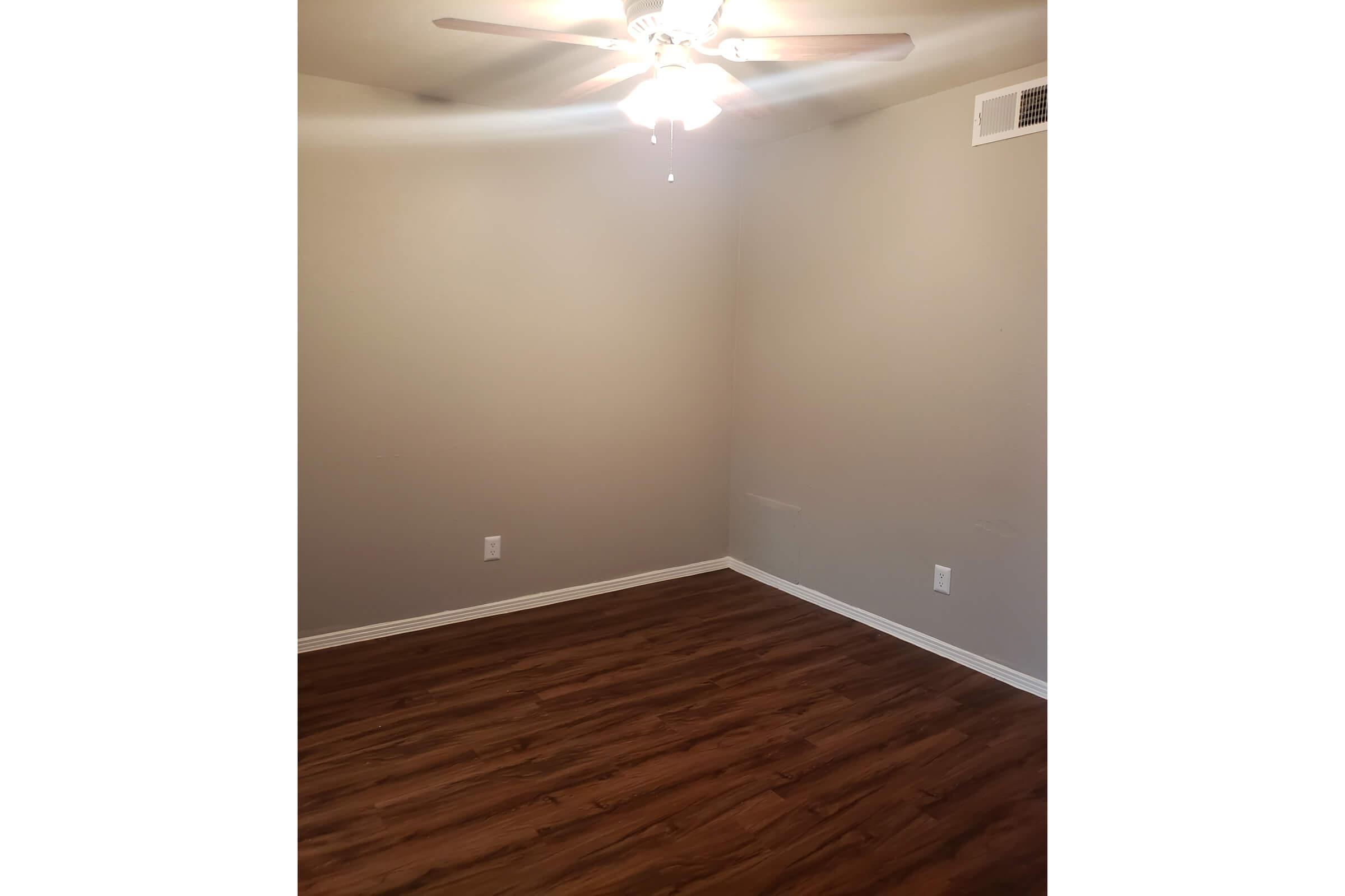 A spacious room with neutral beige walls and a large ceiling fan. The floor is made of light brown wood laminate. There are two electrical outlets visible on the wall. The room is unoccupied and uncluttered, creating a simple and clean aesthetic.