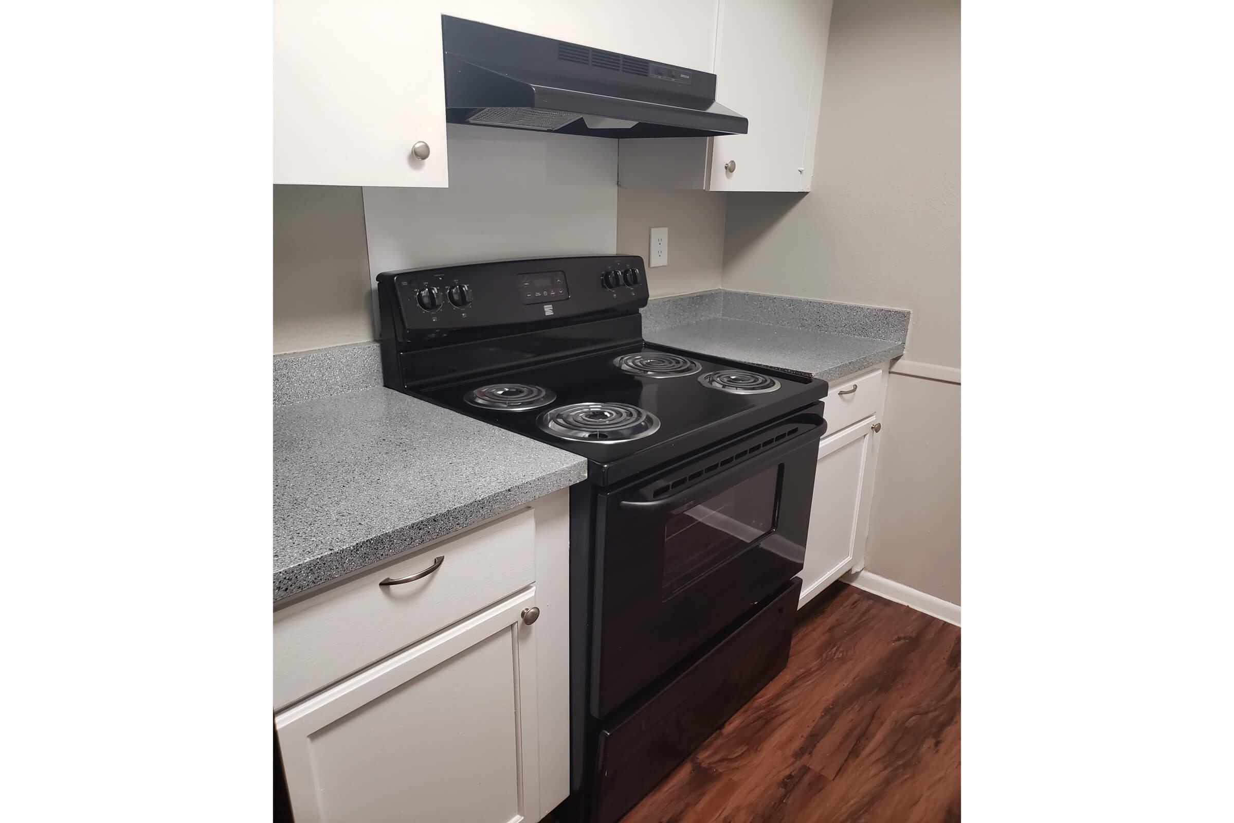 A modern black electric stove with four burners, located in a kitchen with light gray walls and white cabinetry. The stove sits on dark laminate flooring, and there is a range hood above it. Countertops are gray, providing a clean and simple kitchen design.