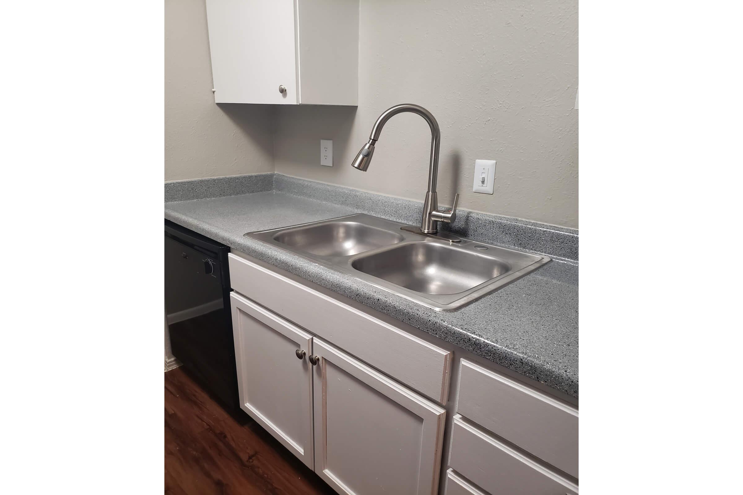 A modern kitchen sink area featuring a double stainless steel sink with a sleek, single-handle faucet. The countertop is gray and the cabinets below are white. The walls are painted a light gray, and there are electrical outlets visible above the countertop. The flooring appears to be a wooden laminate.