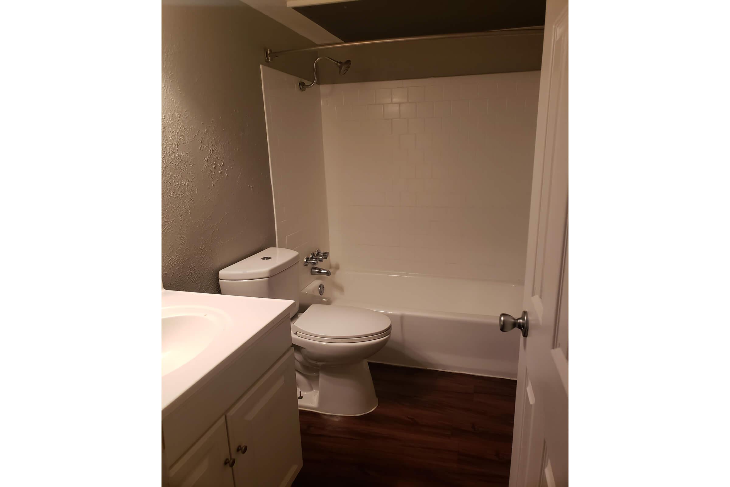 A clean and simple bathroom featuring a white bathtub and shower, a toilet, and a single sink vanity. The walls are painted in a light color, and the floor has a wood-like appearance. The room is well-lit, and the door is partially open, leading into the space.