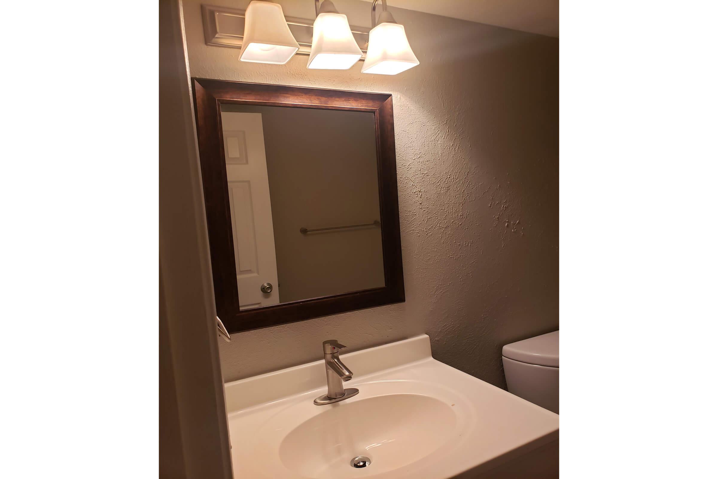 A well-lit bathroom vanity featuring a white sink with a modern faucet, a rectangular mirror framed in dark wood, and three light fixtures above. The wall is a neutral color, and a towel bar is visible in the background, alongside a white toilet.