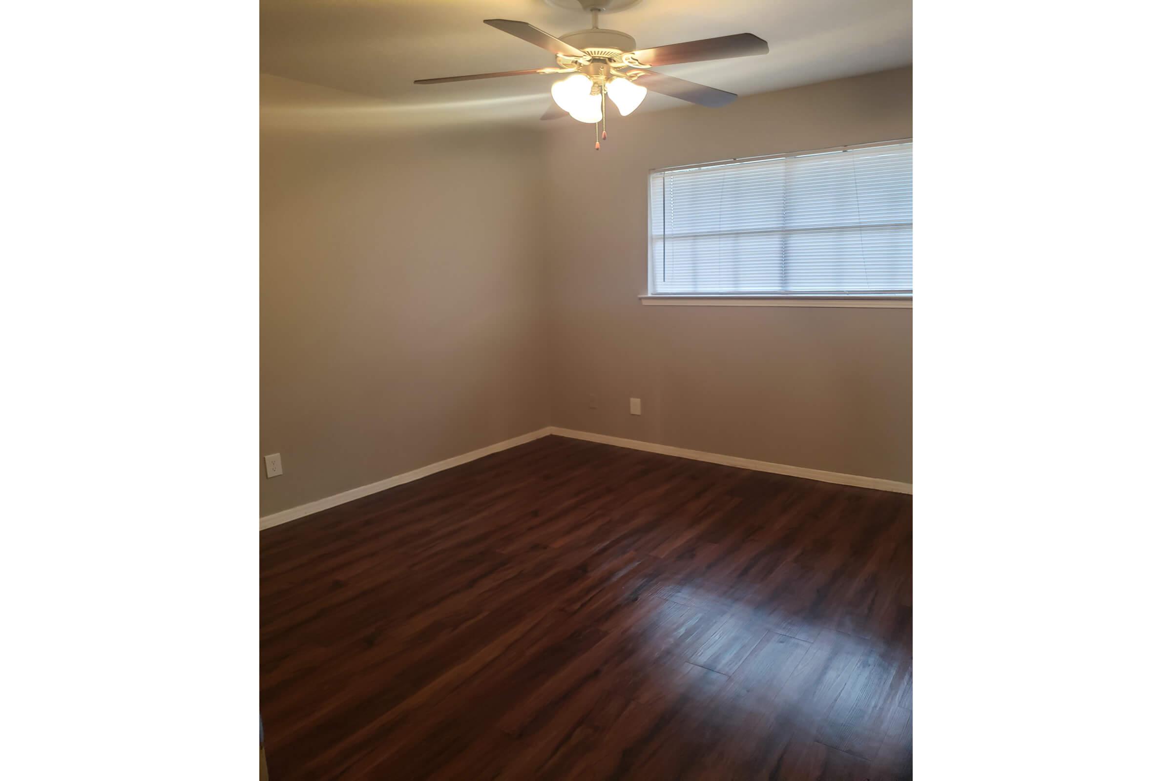 A simple, undecorated room with light gray walls and a ceiling fan. The hardwood floor has a warm finish. Natural light comes in through a window covered with horizontal blinds, casting soft shadows on the floor. The space feels open and minimalistic, ready for personalization.