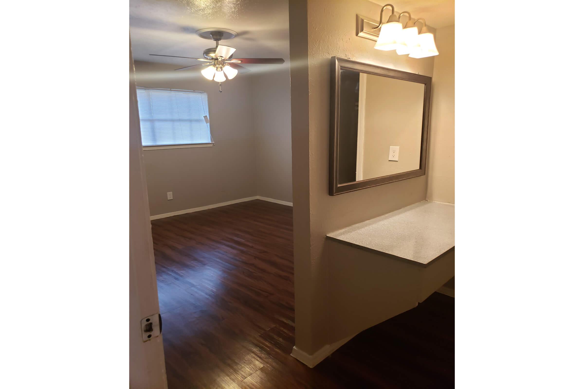 A view of a small, well-lit room with dark wooden flooring. The room features a ceiling fan, a window with blinds, and a wall-mounted mirror. There's a countertop on one side, and the walls are painted in a neutral beige color, creating a warm and inviting atmosphere.