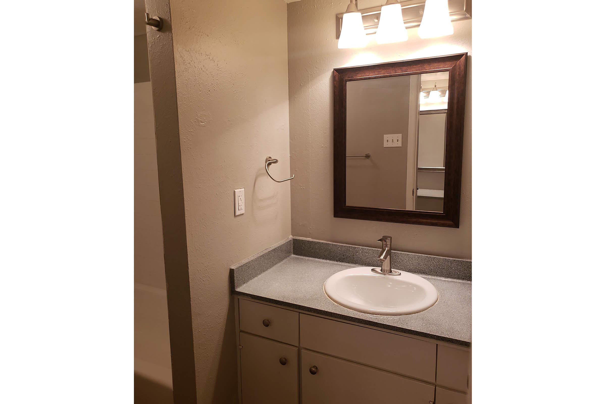 A well-lit bathroom featuring a gray countertop with a sink, a wooden-framed mirror above it, and a towel rack. The walls are painted in a neutral tone, and there is a shower area partially visible in the background. The space appears clean and organized.